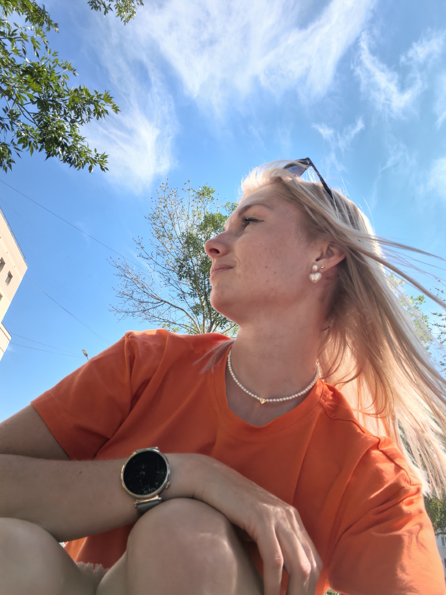 An outdoor, low-angle, medium shot captures a blonde woman from the chest up, looking to her left against a bright blue sky with scattered wispy clouds. She is wearing an orange t-shirt, a pearl necklace with a small heart pendant, and heart-shaped pearl earrings. Her hair is windswept, and sunglasses are perched on her head. On her left wrist, she wears a smartwatch with a black circular face and a gray band. In the background, a portion of a beige building and some green foliage are visible. The scene appears to be a sunny day in the city of Sterlitamak, Russia.