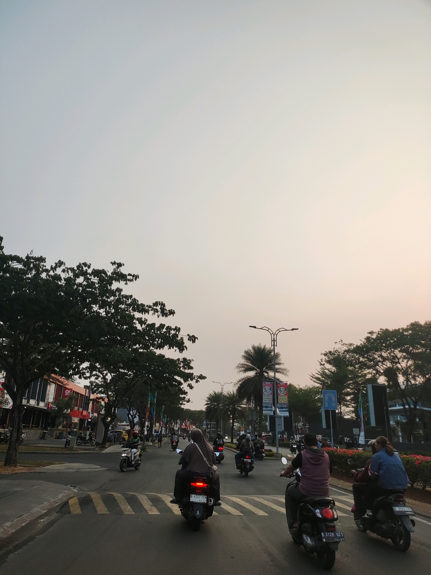 The image depicts a street scene in Telagaasih, Indonesia, under an overcast sky. The foreground shows a road with several motorcycles in motion. On the right side, two individuals are riding a scooter, with the passenger wearing a blue denim jacket and the rider in a maroon hooded shirt. To their left, another motorcycle is driven by a person in a dark hooded garment, and a third motorcycle is further ahead with a single rider. In the middle ground, more motorcycles are visible, including one with a rider wearing a brown hijab and a dark jacket, and another with a rider in a white and green outfit. On the left side of the road, there are buildings with storefronts and trees lining the sidewalk. Palm trees and streetlights are also present. The road surface has yellow and white pedestrian crossing markings. The overall impression is that of a typical urban commute in the late afternoon or early evening.