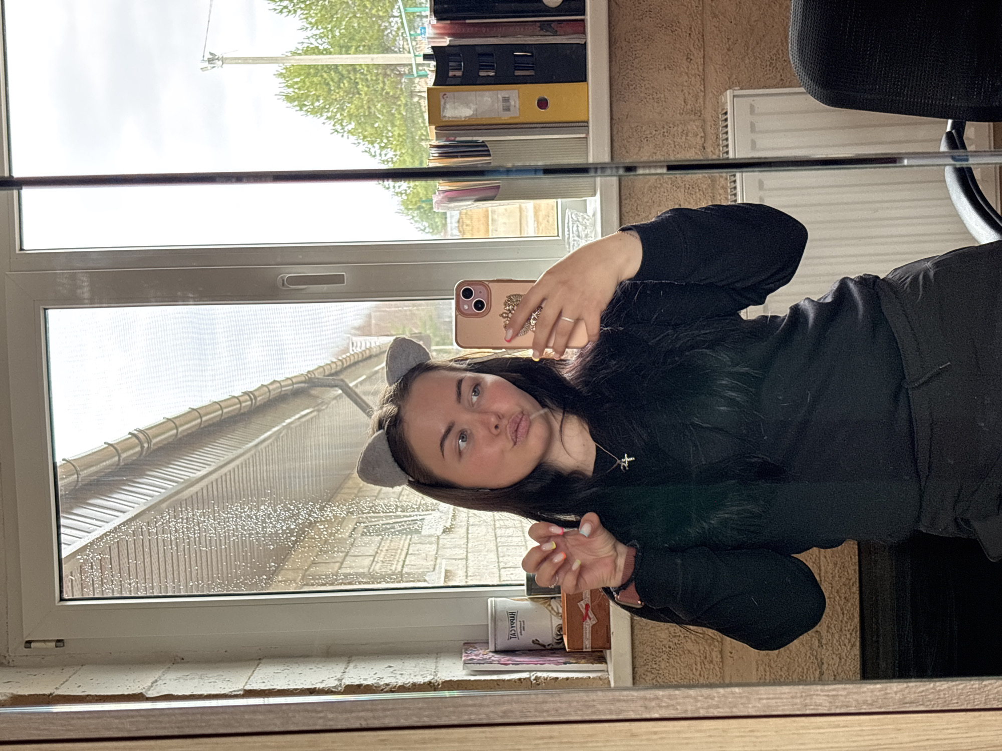 A young woman wearing gray cat ears takes a selfie in front of a window. Raindrops cover the glass, obscuring the view of buildings and a roof. The scene is likely located in Русский Вожой, Russia, given the prompt context. A bookshelf with folders and books is visible behind her, along with a chair and a radiator. Small boxes sit on the windowsill below.