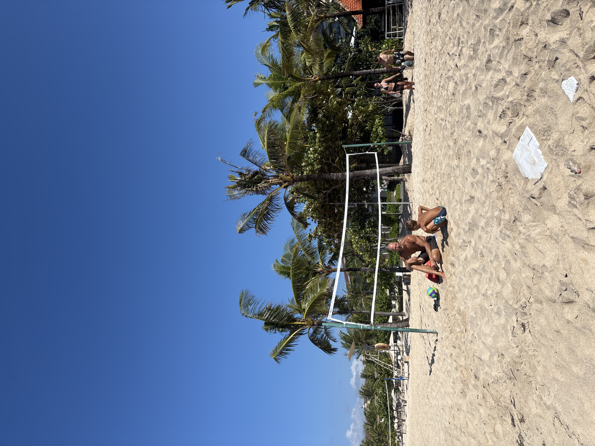 This image captures a bright, sunny day at a beach in Kuta Selatan, Indonesia. The scene is dominated by a sandy expanse, clearly delineated as a casual beach volleyball court by a white net strung between two green poles. Tall, lush palm trees line the background, providing shade and a tropical ambiance, with hints of resort buildings and beach chairs visible behind them.
In the foreground, a shirtless man with a beard, wearing dark shorts, is seated on the sand next to the volleyball net, with a colorful green, purple, and yellow volleyball resting nearby. Partially visible behind him is a young boy in blue and white patterned swim trunks, also on the sand. Further back on the right side of the image, two more people stand on the sand: a man in dark patterned shorts and a woman in a dark bikini top and light bottoms.
The activity appears to be a casual gathering around a beach volleyball setup, possibly before, during, or after a game. The weather is clear and bright, with a vast, unblemished blue sky overhead and strong sunlight casting distinct shadows, suggesting midday or early afternoon. The overall atmosphere is relaxed and recreational. On the sand in the mid-ground, there are a few pieces of white debris and an empty plastic bottle. No discernible text is visible anywhere in the image.