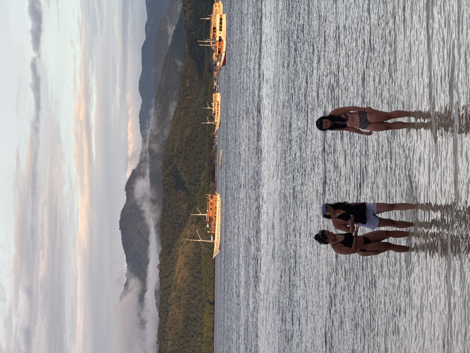 The image shows a coastal scene, possibly Rinca Village, Indonesia, featuring three individuals on a body of water, likely the sea or a large lake. From left to right:
- In the right foreground, a woman in a bikini with a black and white print stands in the water.
- Further down, two women are seen in the water. One wears a black bikini, while the other wears shorts and a dark tank top. The individual in the shorts may be holding a cell phone.
- In the background on the left, there's a forested coastline.
- Multiple ships are anchored near the coastline.
- The sky shows the colors of a rising or setting sun.