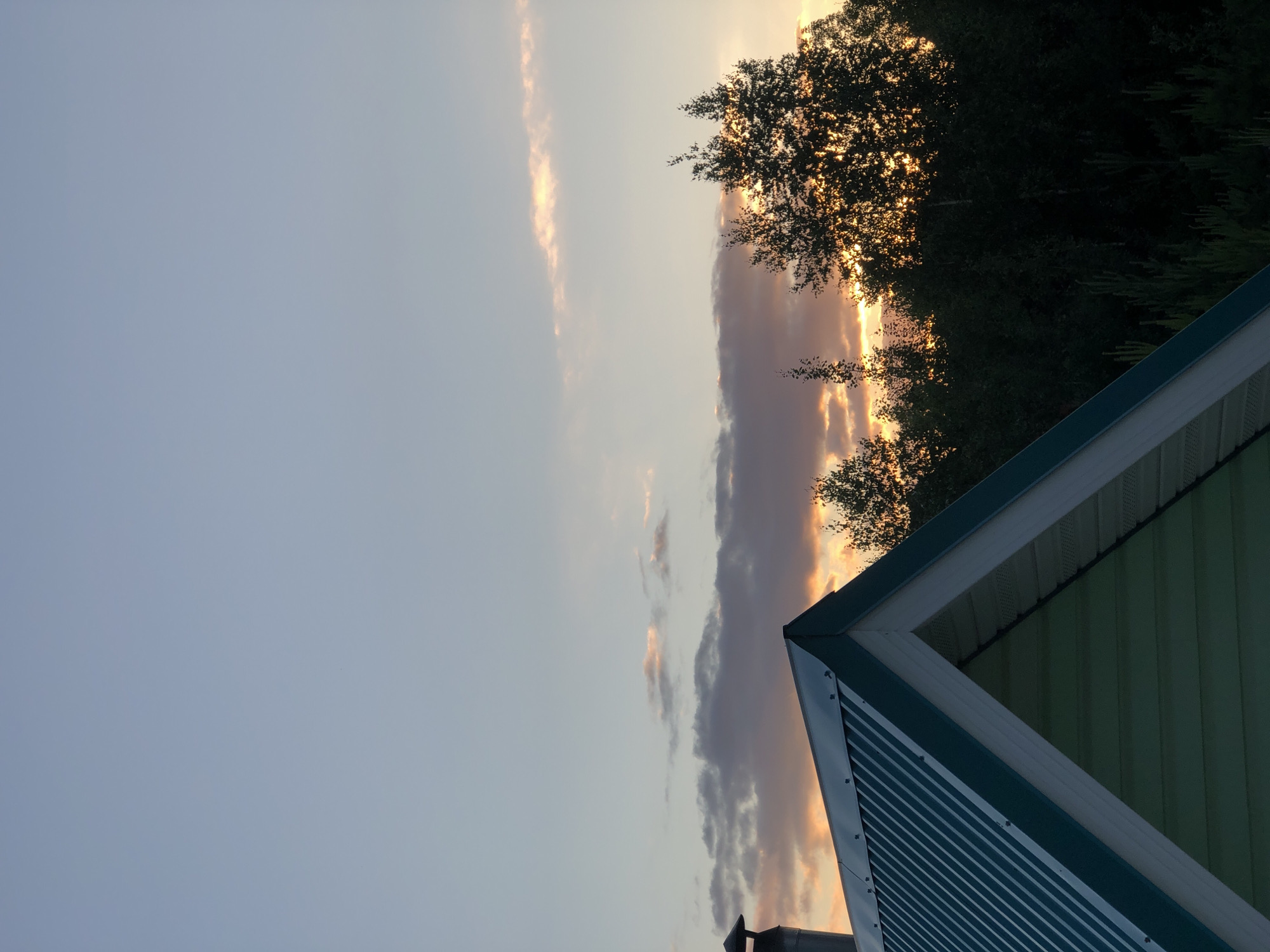 This is a photograph taken at sunset in Noviy, Russia. The image features the roofline of a green-gabled house in the foreground, with a chimney visible on the left side. Behind the house, a dense line of dark green trees is silhouetted against a sky filled with soft, diffused light. Wispy clouds are scattered across the upper portion of the sky, with some tinged with orange and pink from the setting sun. The light filtering through the trees suggests the sun is low on the horizon, casting a warm glow. There are no people or animals visible in the image. The scene depicts a tranquil, natural environment on the outskirts of a settlement.