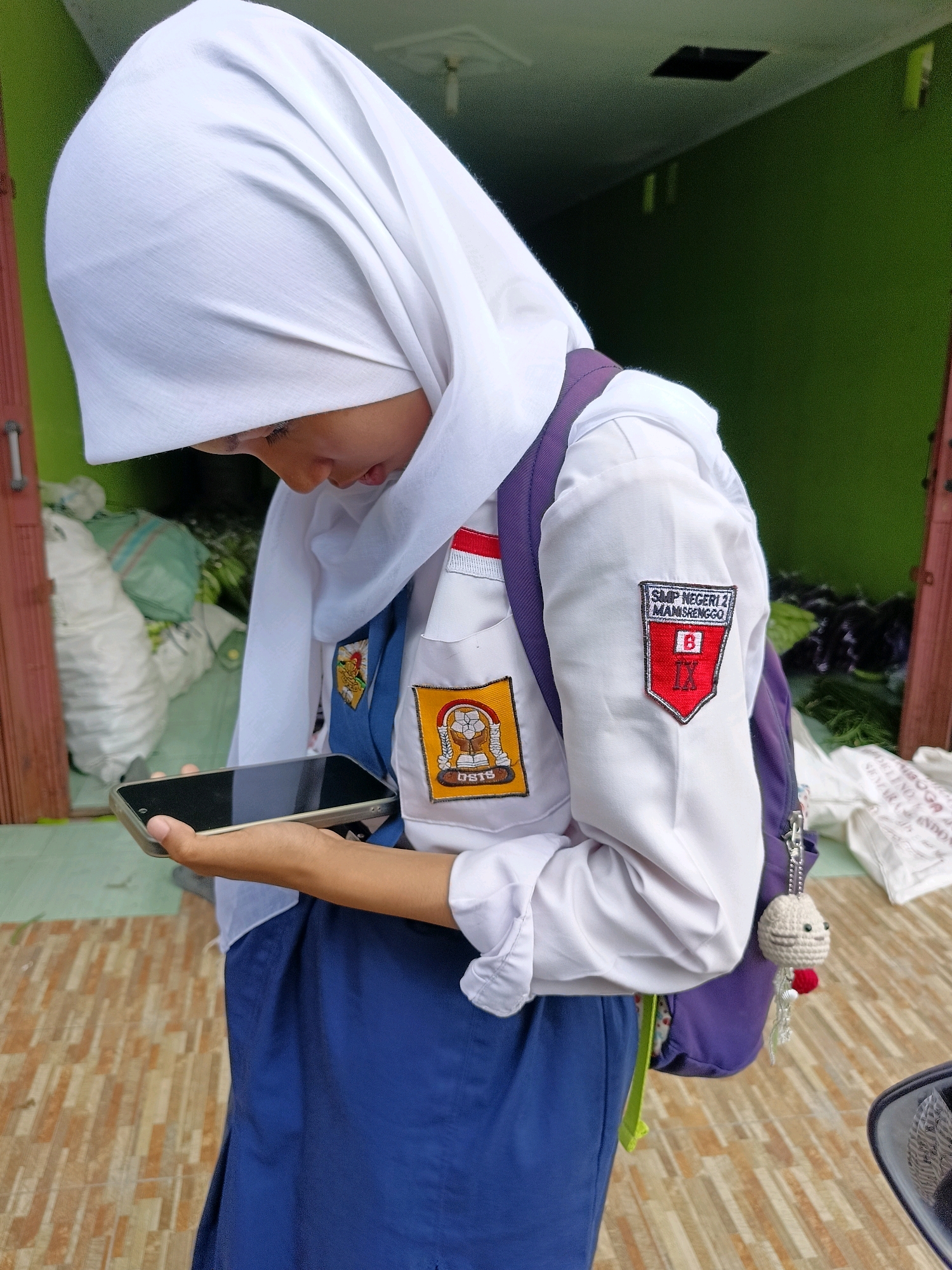 A student in a white hijab and school uniform, which includes a white long-sleeved shirt with patches and a blue skirt, is looking down at a smartphone held in her left hand. The student is standing outdoors, with a green wall and a doorway visible in the background. One patch on the student's shirt reads "SMP NEGERI 2 MANISRENGGO". A purple backpack is slung over her right shoulder, adorned with a crocheted keychain. The ground beneath her is paved with brown and tan tiles. The environment suggests a school or a public area in Manisrenggo, Indonesia.