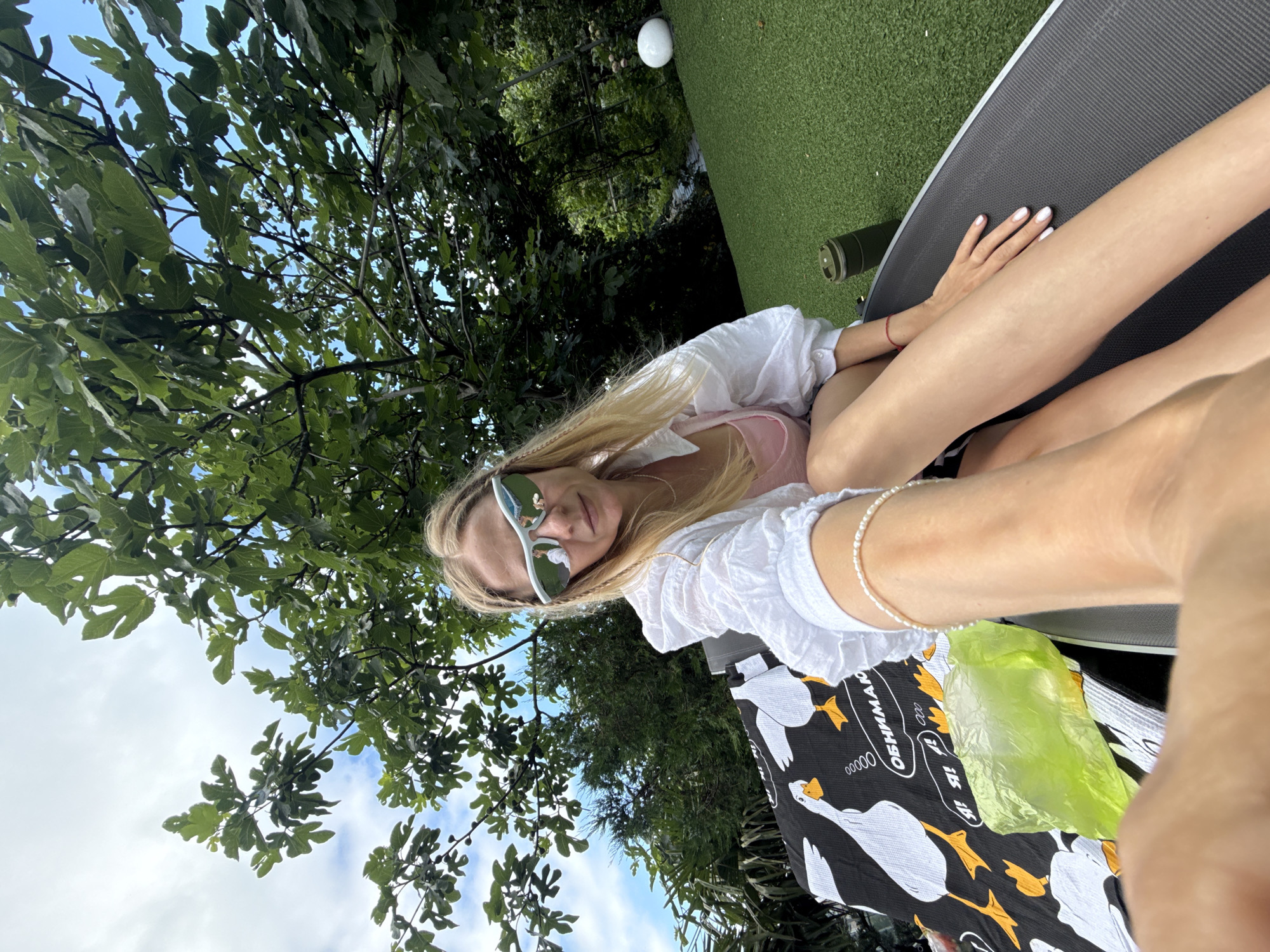 In an outdoor setting, a woman with blonde hair, wearing stylish white sunglasses, a pink top, and a white shirt, sits on a lounge chair. She is facing the camera, with her legs extended forward. A black and white towel with a duck pattern is spread on the chair next to her, and a bright green plastic bag lies on the towel. The background features lush green trees and a bright sky, with artificial turf visible in the distance. A white spherical object rests on the turf. The scene suggests a relaxed moment in a garden or outdoor leisure area.