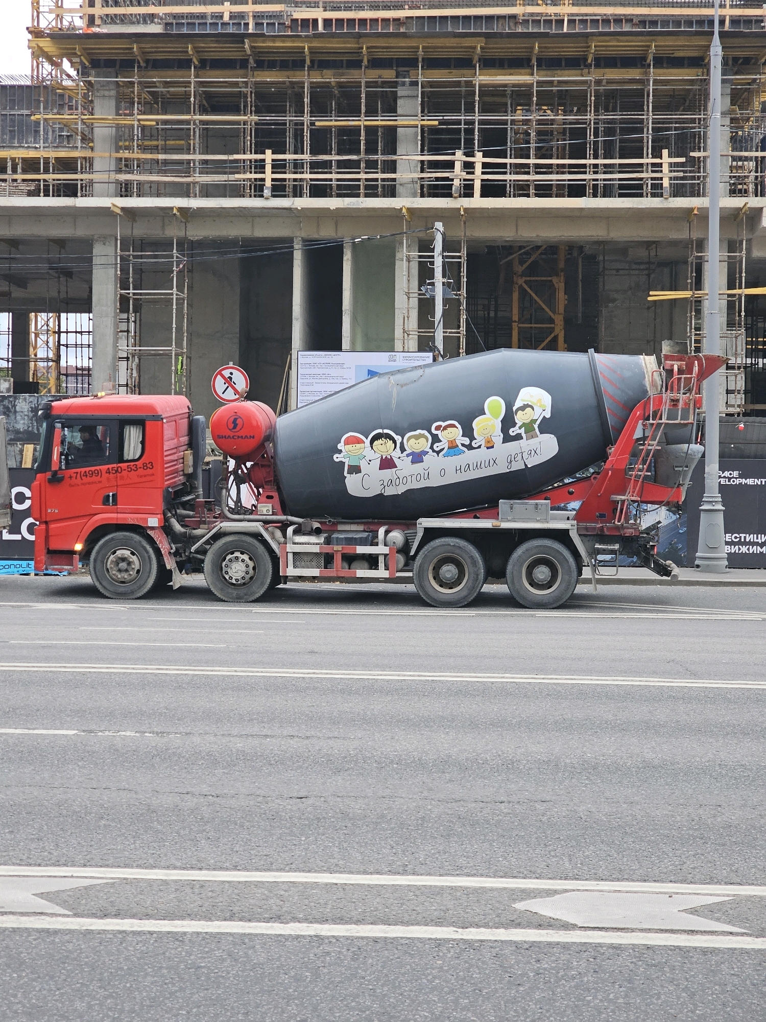 A bright red Shacman concrete mixer truck drives along a paved road in front of a large construction site in Moscow, Russia. The truck has a large, dark gray mixing drum adorned with colorful cartoon stickers of children and a balloon. The truck's cabin is red, and the license plate is not visible. The construction site behind the truck features extensive scaffolding and unfinished concrete structures, indicating ongoing building work. A sign with a red circle and a diagonal line over a person symbol is visible near the construction site, likely indicating a no-entry zone. The overall scene depicts a typical urban construction environment with heavy machinery in operation.