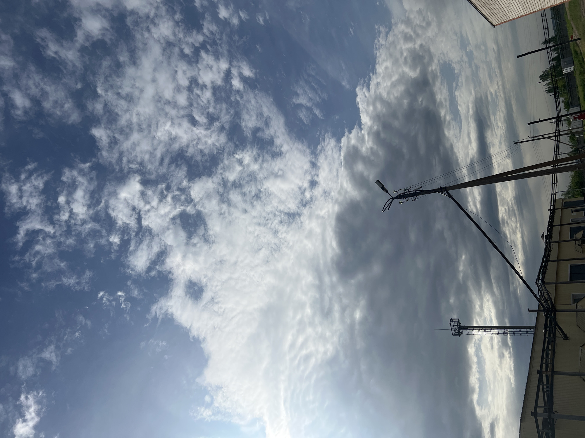 The image shows a daytime view in Бизяки, Russia, looking upwards at the sky. A mix of cirrus and cumulus clouds is visible, creating a varied cloudscape. The sky transitions from a light blue to a darker, more overcast gray. Utility poles and wires extend into the frame from the right side, with a building partially visible in the lower right corner. A transmission tower is also seen among the electrical infrastructure.