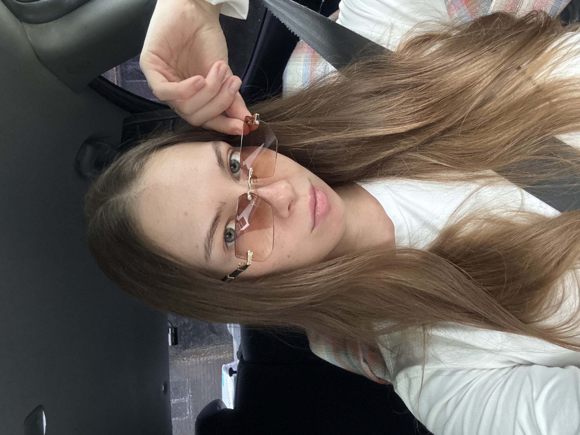 A young Caucasian woman with long, straight, light brown hair is sitting in the passenger seat of a car, looking at the camera. She is wearing a white t-shirt and a colorful plaid scarf draped over her shoulders. She has on rimless, square-shaped sunglasses with peach-colored lenses and gold accents. Her left hand is raised, touching the side of her sunglasses. The interior of the car is visible, including a black seat, a gray seatbelt, and the car's ceiling. Outside the car window, a blurry, overcast sky and what appears to be a brick wall or building can be seen. The lighting suggests it is daytime. This image does not contain any identifiable landmarks or features that would definitively place the location in the city of Perm, Russia.