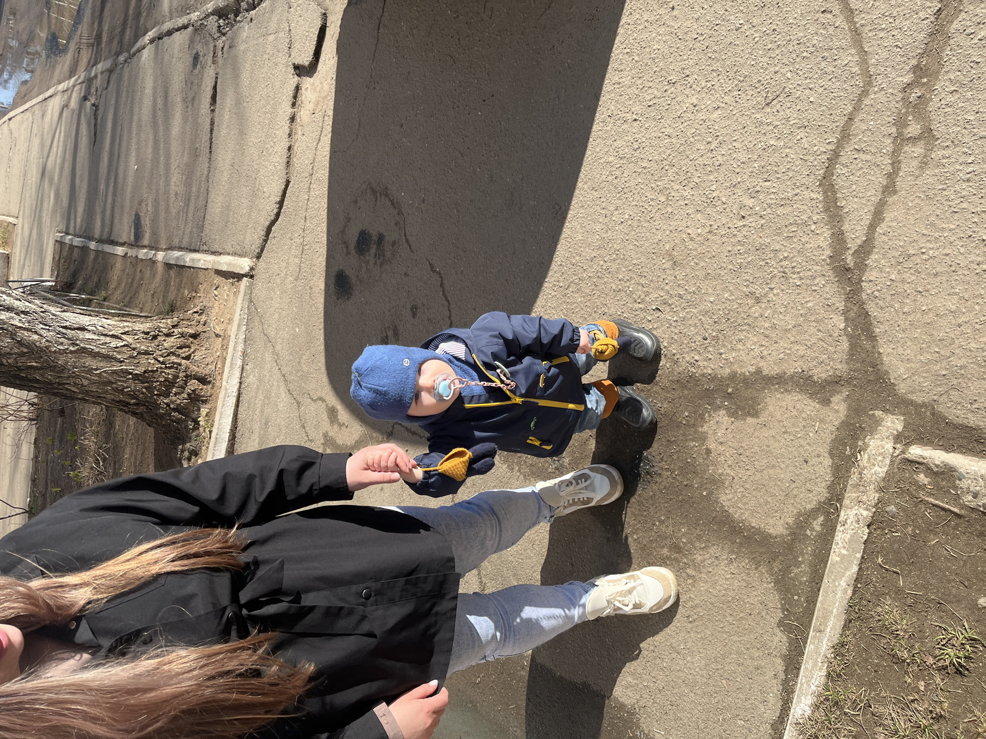 The image depicts a person and a young child on a paved surface, potentially a sidewalk or street in an urban setting. The location is not explicitly identifiable as Chaikovsky, Russia, but the image does not contradict that possibility. The child, wearing a blue hat and dark jacket, has a pacifier and appears to be holding an object (possibly a glove or toy). The person, dressed in a black jacket and light-colored jeans, is holding the child's hand. The environment includes elements like a tree trunk, a concrete structure, and what seems to be an elevated wall, suggesting some landscaping or elevation changes. The light indicates a sunny day.