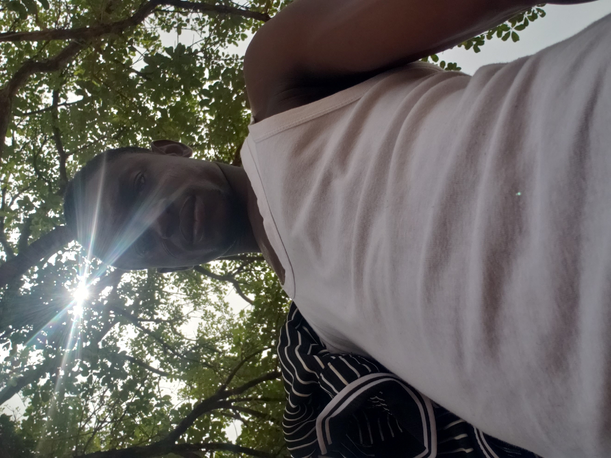 A low-angle, upward-facing shot captures an adult male under a tree. The man, with dark skin, is seen primarily from his face, right shoulder, and upper torso. A bright sun flare, emanating from the upper left, creates a strong starburst effect that partially obscures his face but highlights a subtle smile. He is wearing a white tank top and black-and-white patterned shorts or trousers.
The background consists of dense green tree leaves and branches, silhouetted against a bright, possibly overcast, sky. The intense sunlight and sun flare indicate it is daytime. The image appears to be a selfie taken from a low perspective, with the subject looking down or slightly towards the camera. The setting is outdoors, amidst natural foliage, specifically identified as Adankolo, Nigeria. There is no visible text in the image.