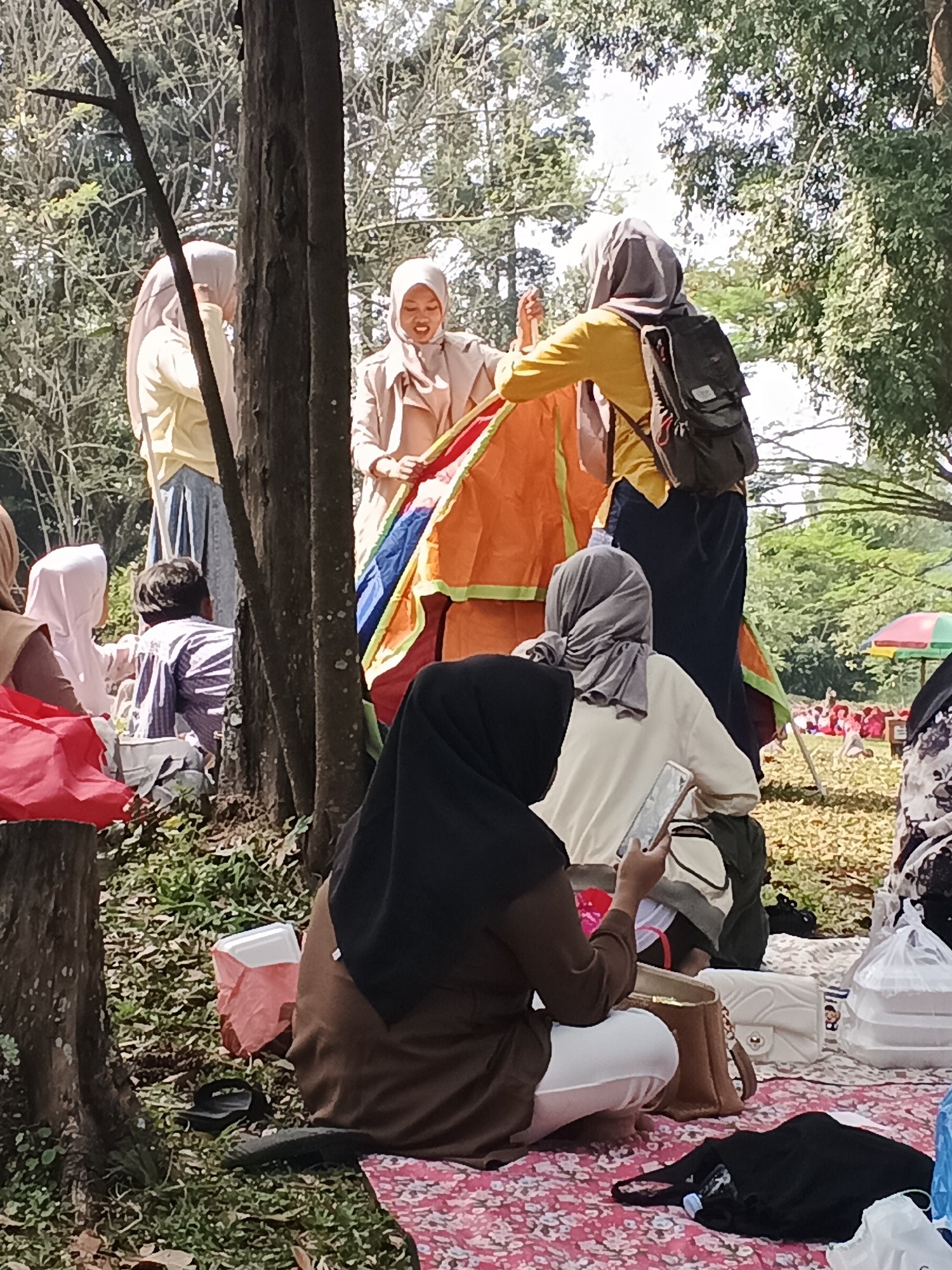 In Perbawati, Indonesia, a group of people are gathered outdoors, likely for a picnic or event in a park. In the foreground, a person wearing a black hijab and brown long-sleeved shirt and white pants is seated, looking at a mobile phone. Next to them is a brown bag and a white purse. Several other bags and items are scattered on a pink patterned blanket.
Behind them, a group of women, some wearing hijabs, are interacting around a large, colorful, tent-like structure that is orange, red, and blue. One woman in a yellow shirt and dark pants is standing, facing away from the camera, wearing a backpack. Another woman in a beige coat is holding a section of the structure. Two other women are partially visible behind them.
In the mid-ground, a person wearing a striped shirt is seated. The environment appears to be a grassy area with trees providing shade, suggesting a natural, park-like setting. In the background, a bright pink and green umbrella is visible, and further in the distance, a group of people are congregated, some wearing red. The overall lighting indicates it is daytime, likely sunny.