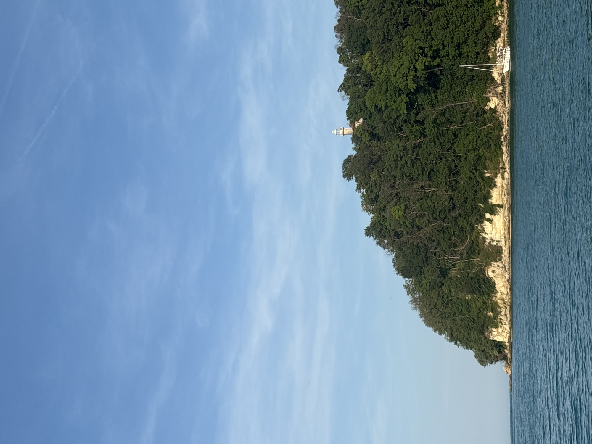 The image captures a coastal landscape under a light blue sky featuring scattered white wispy clouds and a faint contrail in the upper right quadrant. A large, densely forested landmass, appearing as a cliff or a hill, dominates the midground, extending from the left to the right. The trees are primarily dark green deciduous types. The base of the landmass, where it meets the water, consists of a visible light beige rocky cliff face with horizontal striations. Positioned atop the forested elevation, slightly right of center, is a white and beige lighthouse with a distinct domed top. In the lower right midground, a white single-masted sailboat is present in the water, positioned near the base of the cliff, with several figures visible on its deck. The foreground and the left side of the image consist of a body of blue-green water with subtle surface ripples. The scene is illuminated by clear daylight. Location: Unknown, Unknown.