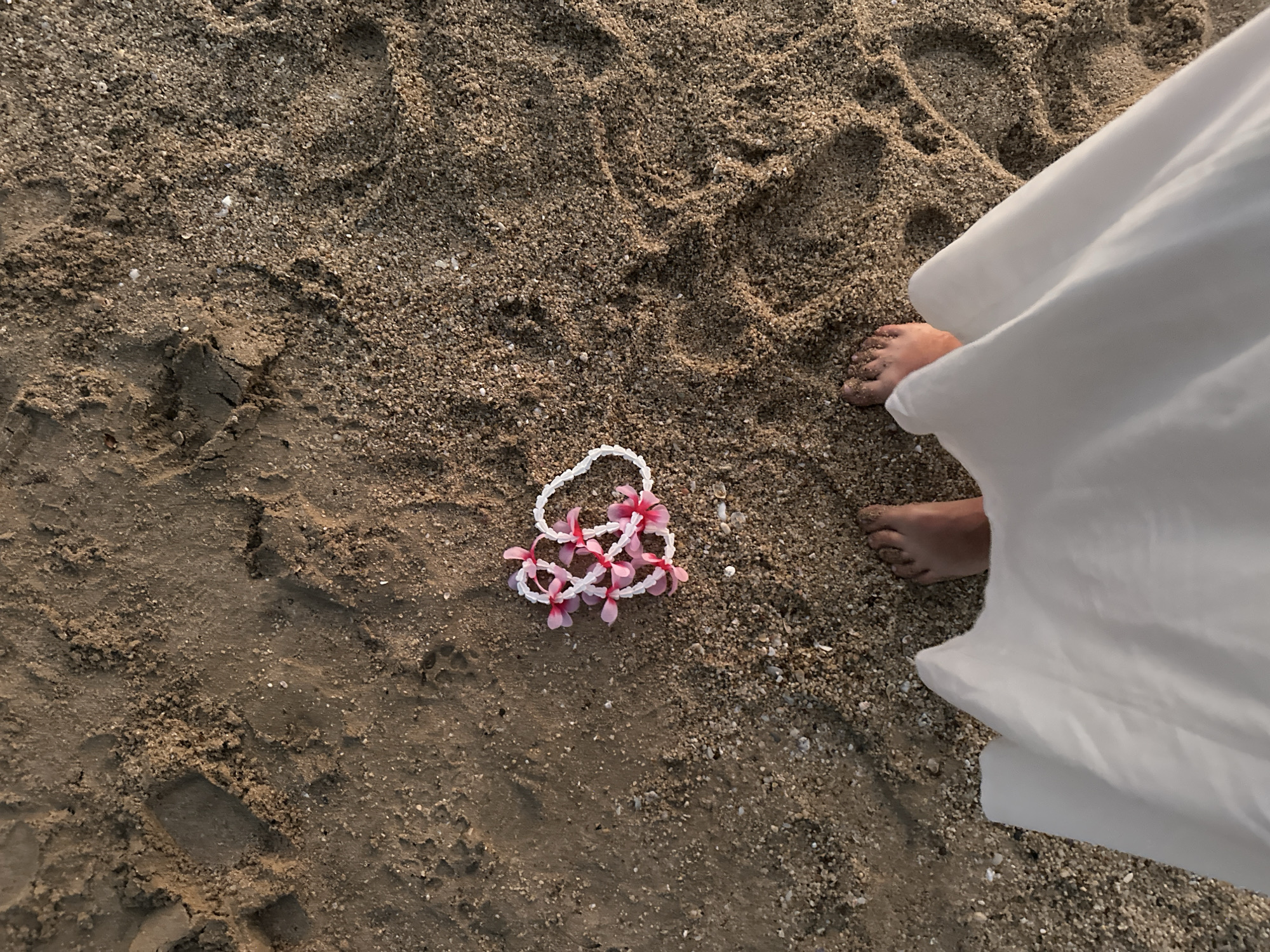 A high-angle shot shows a sandy beach in Pattaya City, Thailand. To the left is a lei of white beads and pink flowers lying on the sand. To the right are a person's feet and a white cloth.
