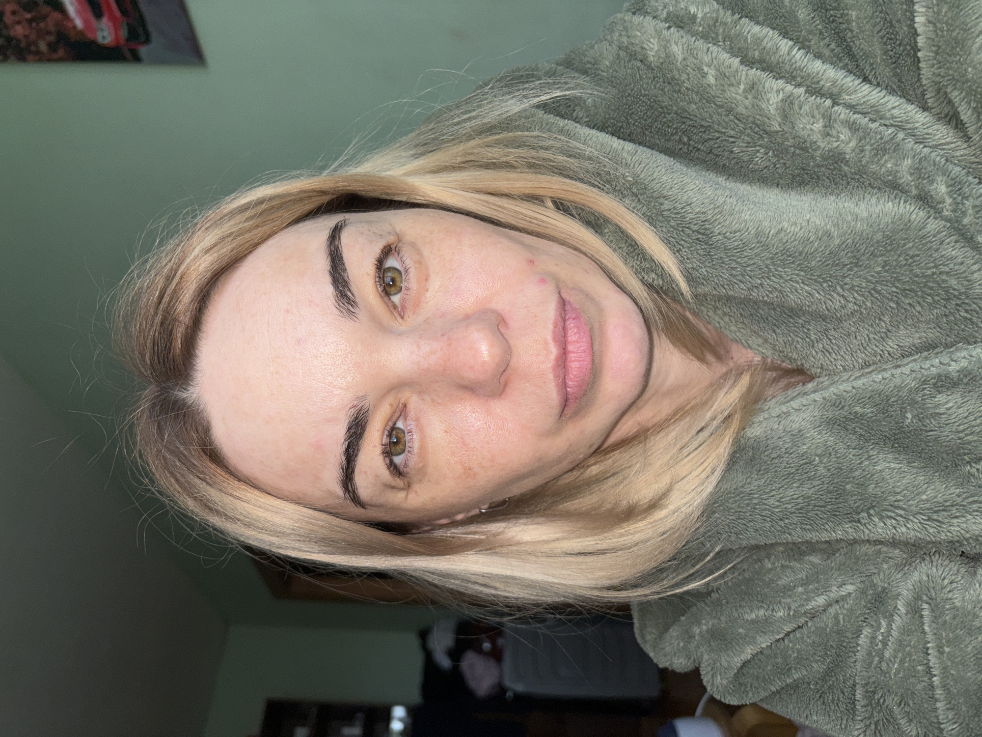 A close-up portrait features a woman looking directly at the viewer with a calm, neutral expression. She has light blonde hair, parted on her left side, with slightly darker roots visible. Her eyes are a striking green-hazel, complemented by dark, well-defined eyebrows. Her fair skin displays numerous freckles across her nose and cheeks, along with a few small red blemishes on her forehead and right cheek. Her lips are a natural pink hue.
She is partially covered by a soft, plush, green-grey robe or blanket, which shows a distinct fuzzy texture. The background consists of a plain green-teal wall. In the upper left corner, a blurred, indistinct object that appears to be a picture frame with a red item (possibly a car) inside is visible. Other dark, blurry shapes in the lower background suggest an indoor setting, likely a casual home environment. The lighting is soft and even, indicating an interior space. There is no visible text in the image.