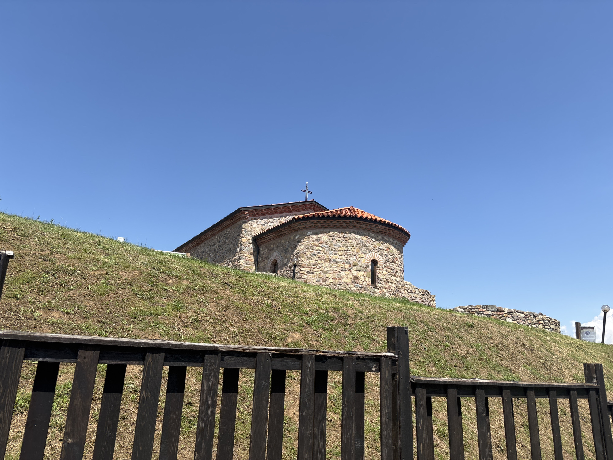The image displays a stone ecclesiastical building, likely a church or monastery, positioned on a grassy hill under a cloudless blue sky. The structure, located in Belchinski Bani, Bulgaria, is constructed from natural, irregularly shaped stones and features a distinctive red tiled roof. A dark cross is mounted at the peak of the roof. Several small, arched windows are visible on the building's rounded walls. In the foreground, a dark brown or black wooden fence composed of vertical slats spans the width of the frame. The hillside is covered with sparse green and dry brown grass, revealing patches of earth. To the far right on the hill, a section of a stone wall, a white informational sign, and the top of a lamppost are partially visible. No people are present in the scene.