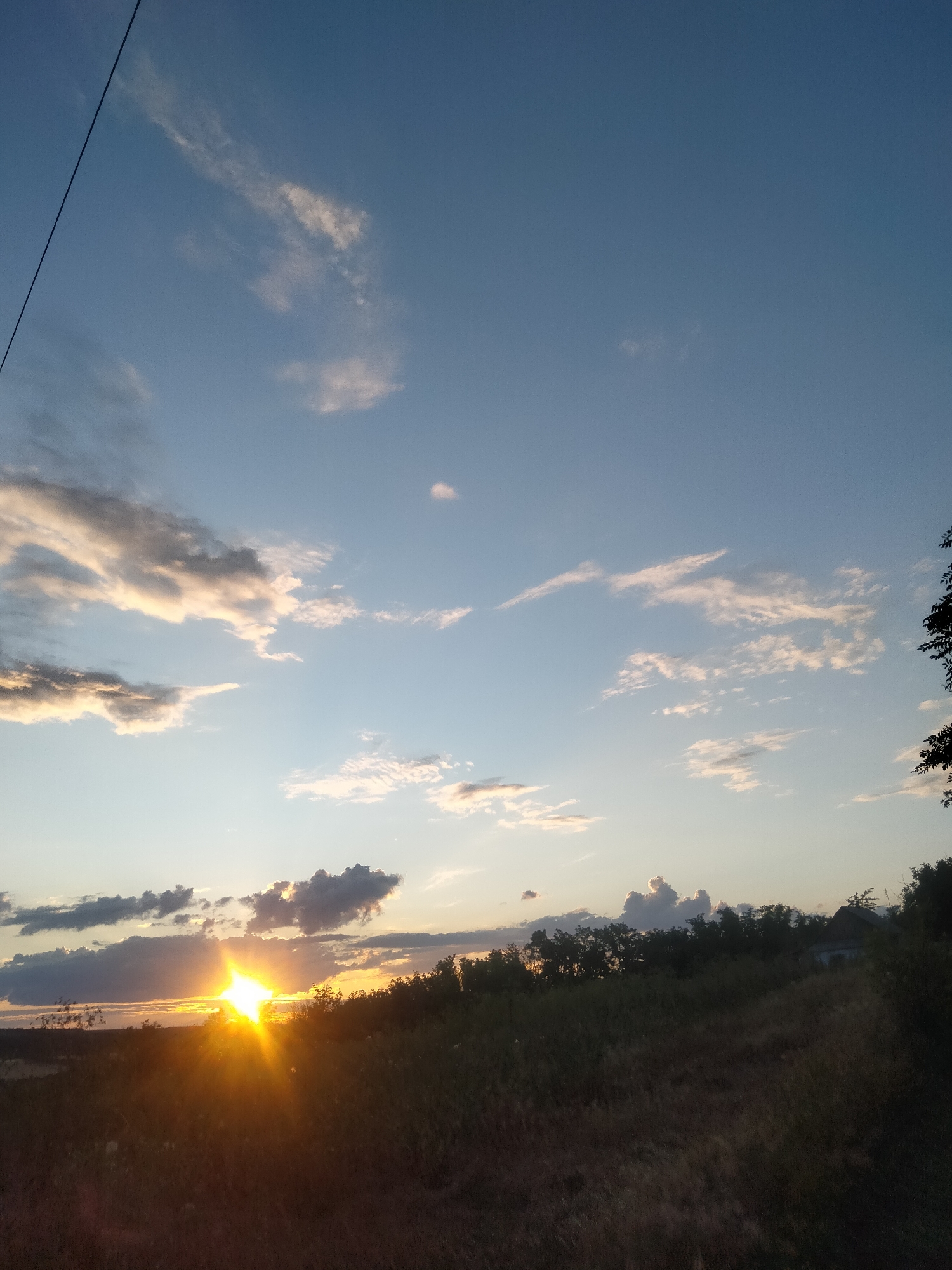 The image depicts a rural landscape at sunset. The sky is a clear blue with scattered clouds, some of which are illuminated by the setting sun, casting a warm glow. The sun itself is a bright, starburst-like shape peeking over the horizon, its rays spreading across the sky. In the foreground, a grassy field slopes upwards towards a line of trees and vegetation. A dark silhouette of trees runs across the middle ground, with a small, dilapidated building visible among them on the right side. A power line stretches diagonally across the upper left portion of the frame. The overall impression is one of a peaceful, natural scene in a rural setting. There are no people or animals visible in the image.