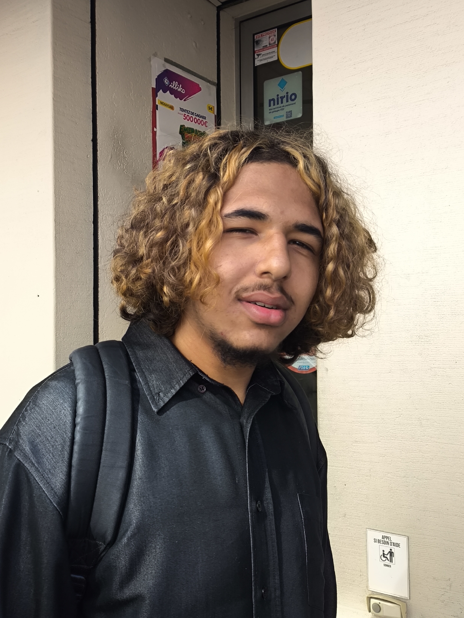 A young man with curly, light brown hair, a short beard, and a mustache is depicted in a portrait-style shot. He is wearing a dark, possibly black, button-up shirt and a black backpack over his left shoulder. He looks directly at the viewer with a slight smirk and a squint in his right eye, suggesting a playful or confident expression.
The background consists of a light-colored wall or door, which appears to be part of a building entrance in Schiltigheim, France, given the contextual information. Several signs are affixed to the wall and door. Visible text includes "SITE PROTEGE" (Protected Site), and a lottery advertisement for "illiko" stating "NOUVEAU TENTEZ DE GAGNER 500000€" (New, Try to win 500,000€). Another sign displays "nirio Vos services de paiement du groupe FDJ" (Nirio, Your payment services from the FDJ group), indicating a payment service associated with the French national lottery. Below, an accessibility sign reads "APPEL SI BESOIN D'AIDE SONNER" (Call if you need help, Ring), next to a white call button. The lighting is soft, suggesting an indoor setting or an overcast day, with no harsh shadows.