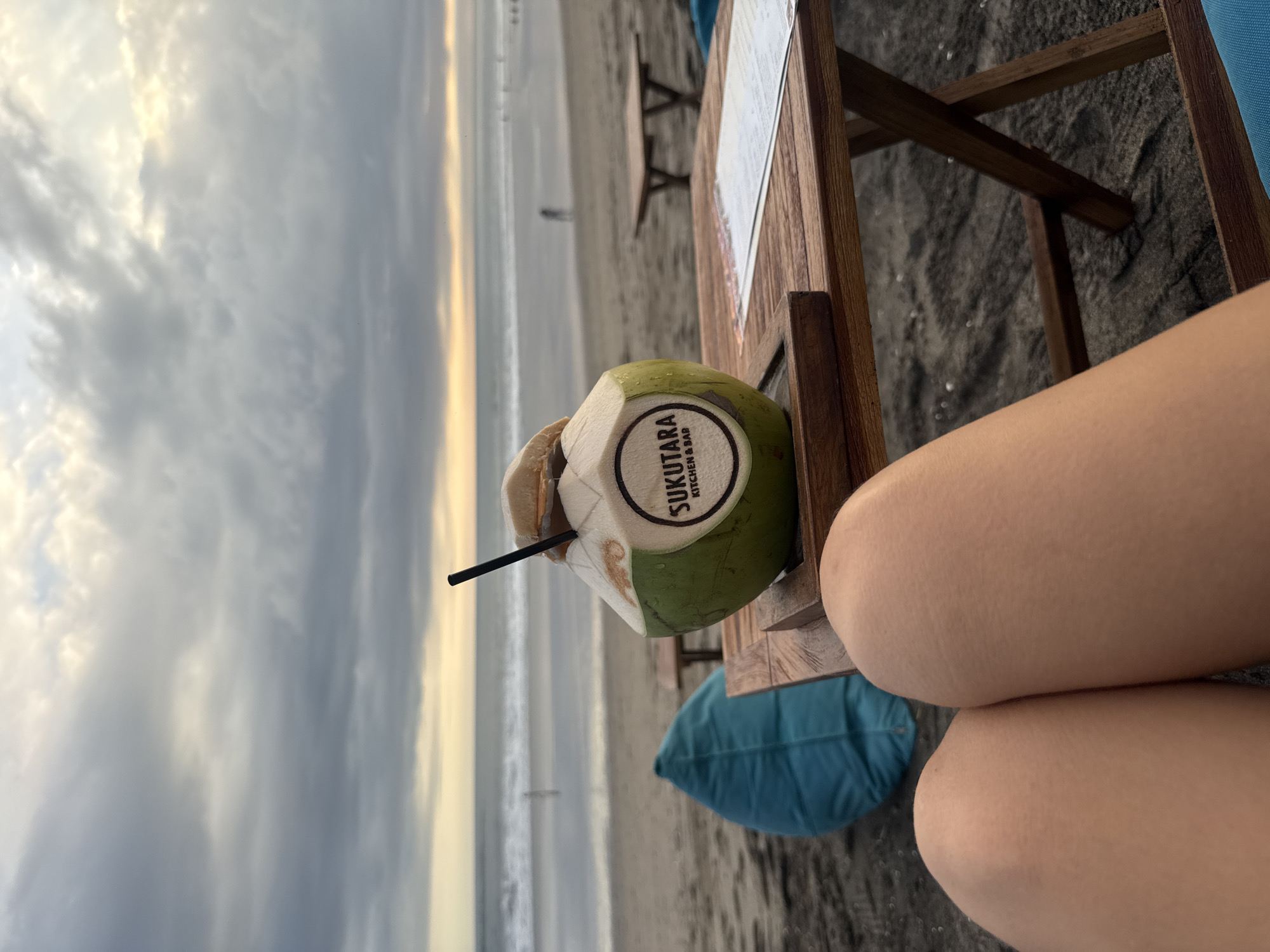 A view from above the beach, a cloudy, almost sunset-tinged sky, and the ocean are visible in the background. A partially visible person, wearing shorts, sits at a rough wooden table on a beach in North Kuta, Indonesia. On the table is a young coconut with a black straw in it. The coconut has a sticker on it with the words "SUKUTARA KITCHEN & BAR" in a round logo. A menu lays partially visible on the table, as well. A blue cushion is on the beach under the table. Other tables can be seen in the distance on the beach.