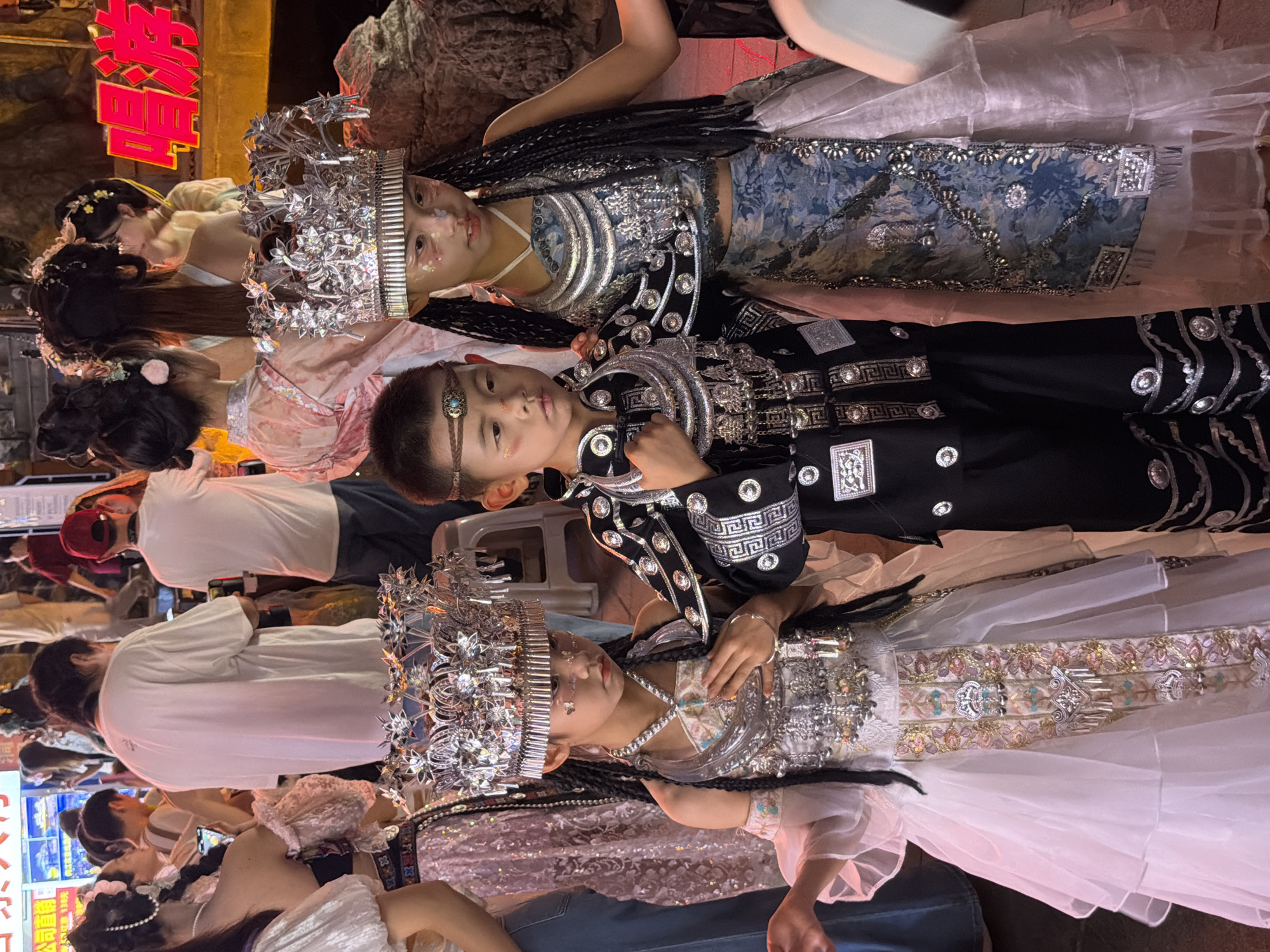 This media file shows a group of people, likely performers, dressed in elaborate, ornate costumes. The subjects are primarily children and young adults, adorned with intricate silver headdresses resembling crowns and adorned with metallic floral motifs and jewels. Their costumes feature a blend of historical and fantastical elements, with rich fabrics, metallic embellishments, and braided hairstyles. One child wears a black outfit with silver geometric patterns and circular ornaments, while others are in lighter, flowing garments. The overall aesthetic suggests a performance or parade, possibly a cultural or festive event.
The scene appears to be an outdoor setting, illuminated by artificial lighting, indicating it could be evening or nighttime. The background includes some rocky textures and a glimpse of a building with neon signage. The people are positioned in a way that suggests they are either posing for the camera or moving as part of a procession. The children's faces show a range of expressions, some looking directly at the camera with a neutral or slightly playful gaze, while others appear more serene or focused. The presence of onlookers in the background, some wearing casual clothing, further supports the idea of a public event.
Notable details include the high level of craftsmanship in the costumes and headdresses, the use of glitter on the faces of some performers, and the general celebratory atmosphere implied by the attire and lighting. A red neon sign with Chinese characters is visible in the upper left corner, with the characters "梦幻" (Mènghuàn - Dreamlike/Fantasy) clearly legible. The location context provided is Yuzhong District, China, which, combined with the visible text, suggests the event is taking place in a populated urban area within China.