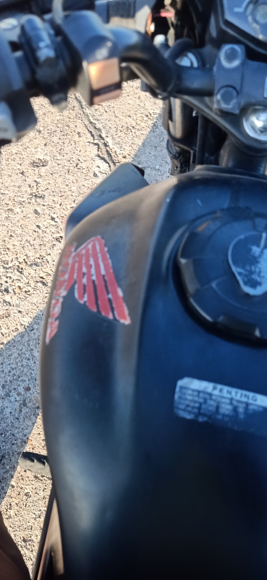 The image shows a close-up of a motorcycle fuel tank, with a red and white Honda logo visible on the left side. The tank is dark blue and appears slightly weathered. A sticker with the word "PENTING" (which means "important" in Indonesian) is present on the lower right of the tank. The motorcycle is parked on a textured surface that appears to be concrete or asphalt, with some gravel. The background is blurred, but suggests an outdoor environment, possibly a roadside or parking area in Keudekruenggeukueh, Indonesia. No people are visible in the image, and no actions are taking place.