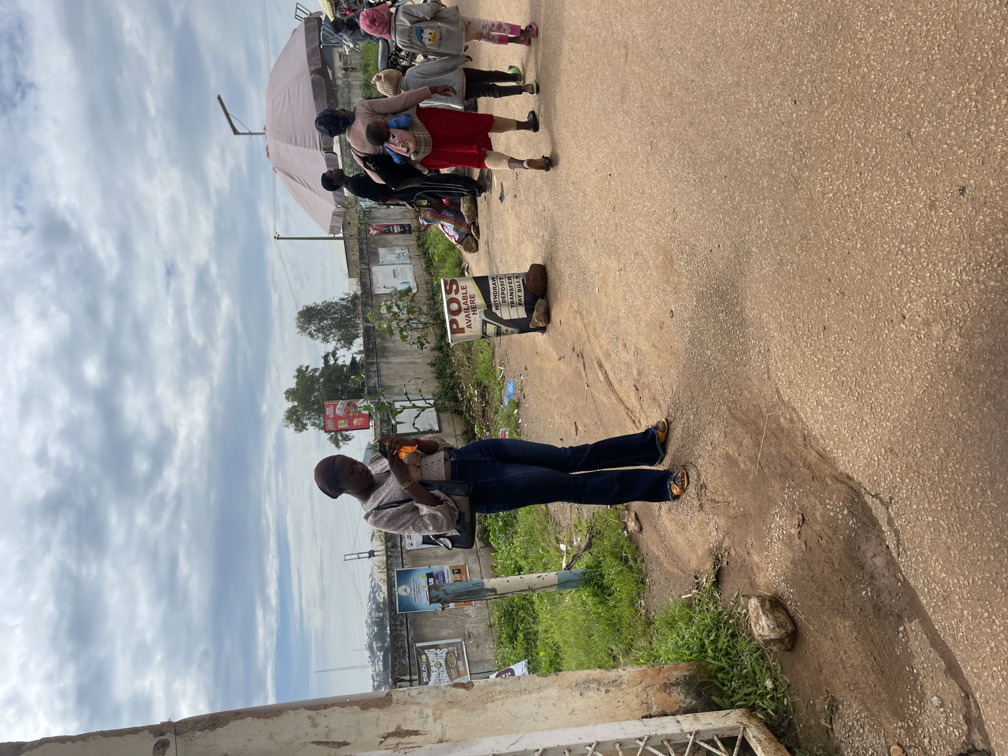 In Rafin Par, Nigeria, a young woman stands on a dirt road, looking down at her orange-colored smartphone. She wears a light-colored, patterned long-sleeved blouse and dark flared jeans, with yellow sandals on her feet. Behind her, a group of people are walking away from the camera. A woman in a red dress carries a baby on her back, and two children, one wearing a grey jacket with cartoon characters and a pink hat, and another in a light-colored hat and grey jacket, walk beside her. A large pink umbrella, possibly for shade, is partially visible behind the woman with the children. To the left of the woman with the phone, a white-colored fence post stands near a wall with posters. A sign on a stand reads "POS AVAILABLE HERE" with options for "WITHDRAW," "DEPOSIT," "TRANSFER," and "PAY BILLS." The ground is a mix of dirt and gravel, with some sparse green grass growing along the side of the road. The sky is overcast with a cloudy pattern.