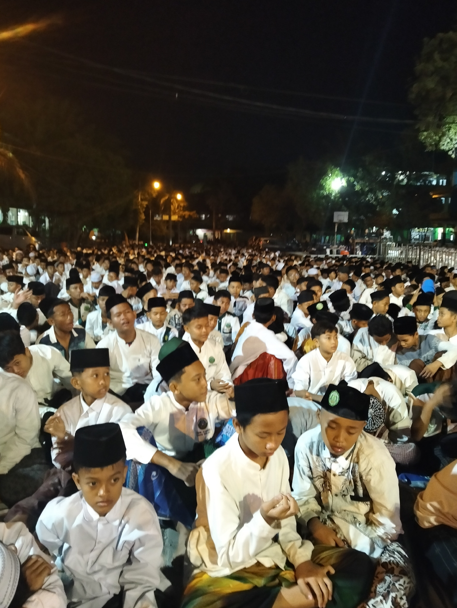 The image shows a large gathering of young males, likely students, seated on the ground outdoors at night. They are all wearing white shirts and black peci hats, common attire for religious events in Indonesia. Many are looking in the same direction, suggesting they are listening to a speaker or watching a performance. The scene is illuminated by artificial lights, and trees and buildings are visible in the background. The image is likely taken at a pesantren (Islamic boarding school) or a religious gathering in Drajat, Indonesia. The atmosphere appears to be one of solemnity and community.