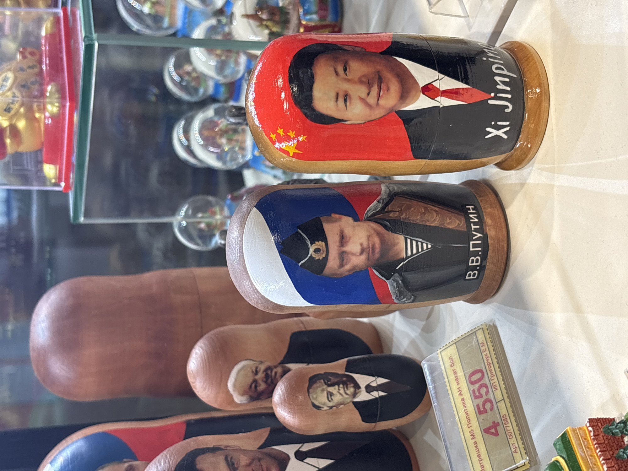 This photograph shows a display of Russian nesting dolls, also known as Matryoshka dolls, with the images of political figures on them. Two dolls are prominently featured in the foreground. The top doll is painted with the image of Xi Jinping, the General Secretary of the Chinese Communist Party, with the name "Xi Jinpin" written in white on the side. The doll below depicts Vladimir Putin, the President of Russia, with "В.В.Путин" (V.V. Putin) written in Russian on the side.
Behind these, a partially visible series of dolls featuring various political leaders are seen. A price tag is also visible on the right side of the dolls. The city and location are unknown.