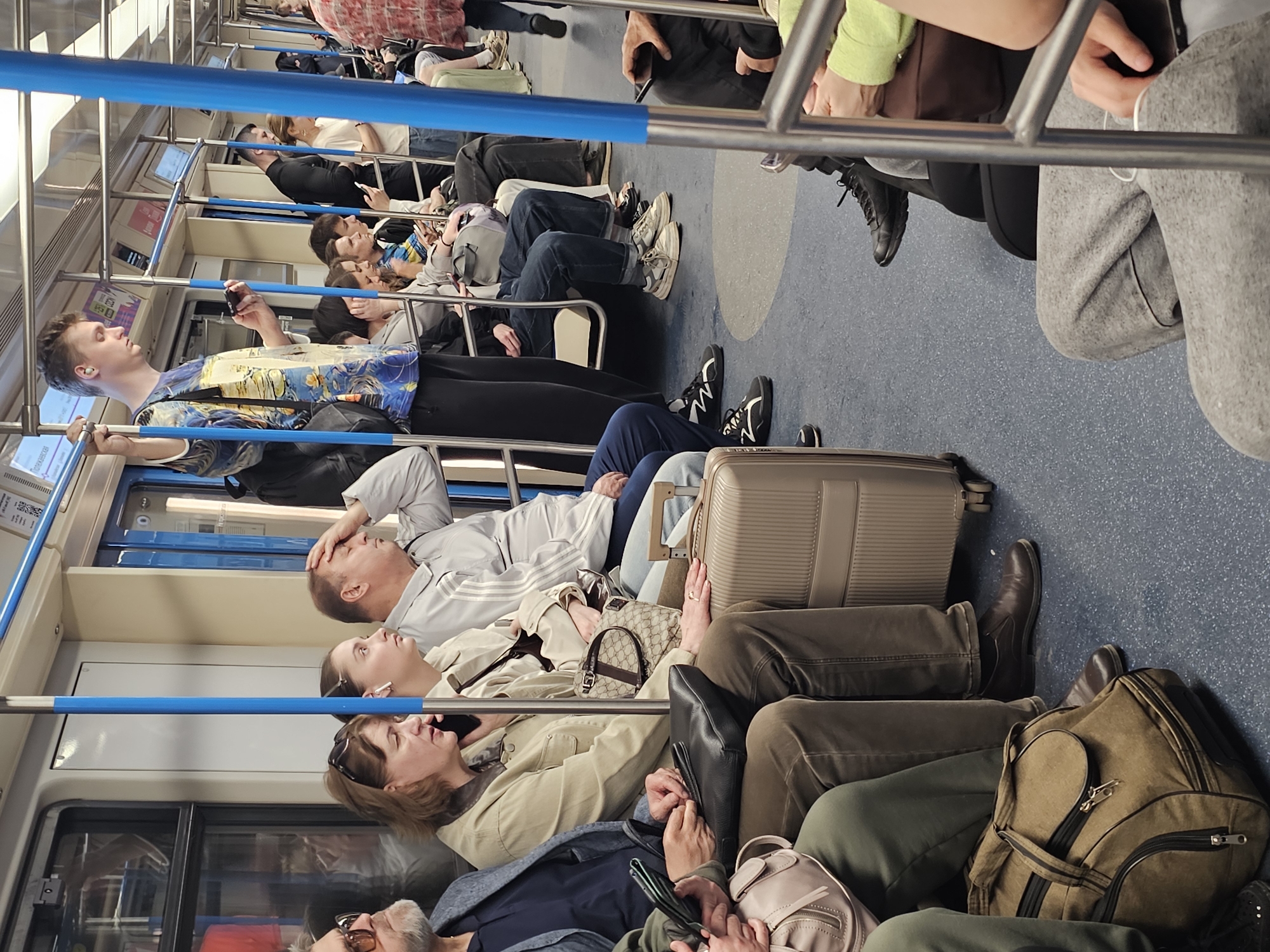 The image depicts the interior of a train, potentially a subway or metro, crowded with passengers. The train is identified as located in Khimki, Russia based on the signage visible. Most passengers are seated and dressed in casual attire. Several are using mobile phones. There are various bags and luggage items present on the floor. The interior of the train appears modern, with bright lighting and a light-colored interior.