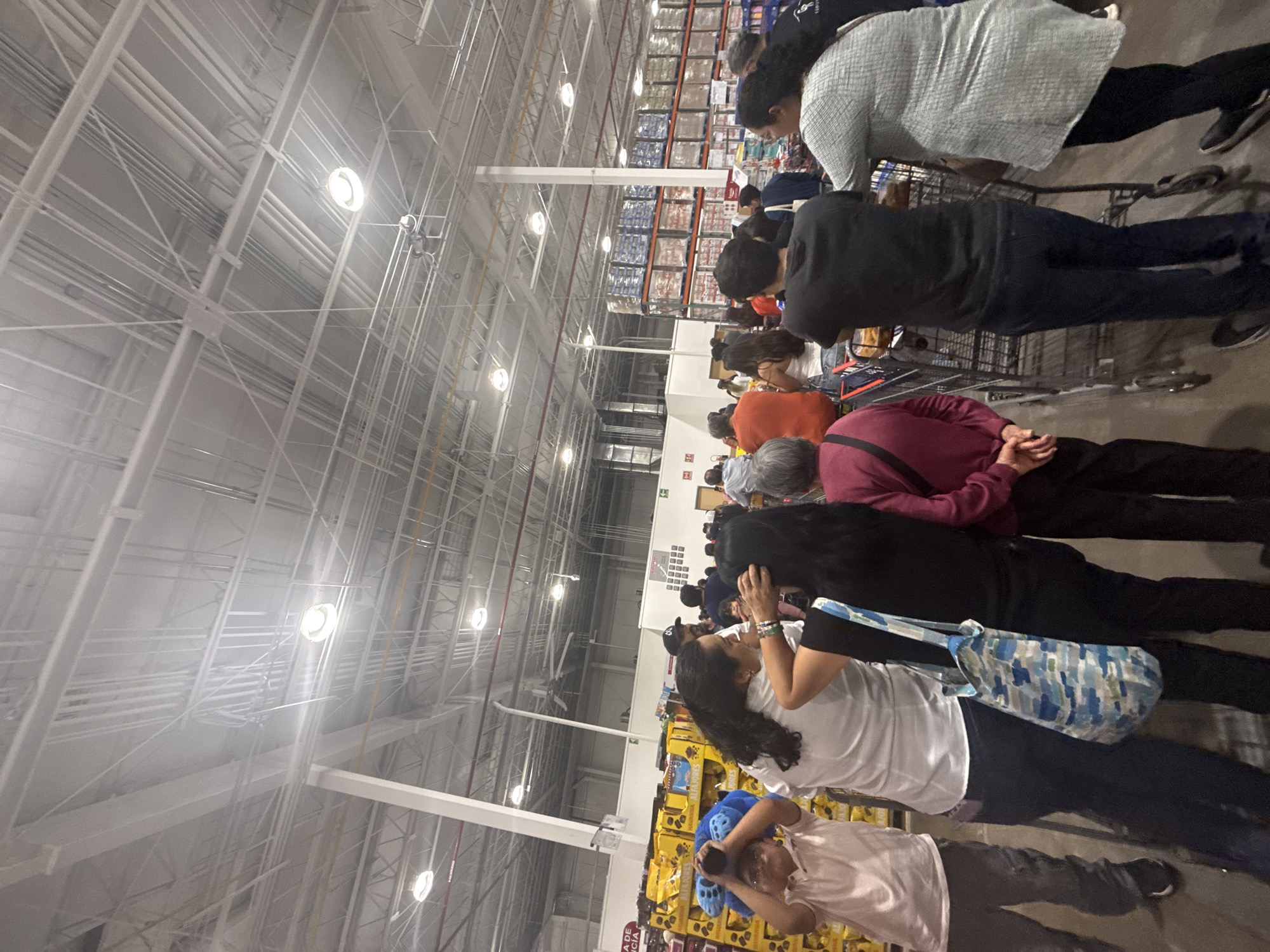 This vertical image depicts a long queue of people inside a large, warehouse-style retail store in Puebla, Mexico. The setting is characterized by a high industrial ceiling with exposed metal trusses, pipes, and bright circular fluorescent light fixtures. Tall metal shelving units, heavily stocked with bulk merchandise in plastic wrap and cardboard boxes, extend into the background on both sides of the aisle where the customers are waiting.
The primary subjects are numerous individuals, primarily adults, forming a queue. Many are standing behind shopping carts, suggesting they are either waiting to check out or for a service within the store. In the foreground, a child wears a distinctive blue, possibly animal-themed, fuzzy hat, and an adult carries a blue and white patterned shoulder bag. Most people are seen from behind or the side, facing forward in the line.
The activity taking place is people waiting in an orderly fashion in a busy retail environment. The time of day is not discernible due to the indoor artificial lighting. There are no visible cues regarding weather. The general atmosphere appears busy but calm, with people patiently standing in line. Visible text is scarce; a partially obscured yellow sign in the foreground shows fragments like "ADE" and "CIA", and some faint text is visible on distant product packaging, but it is unreadable.