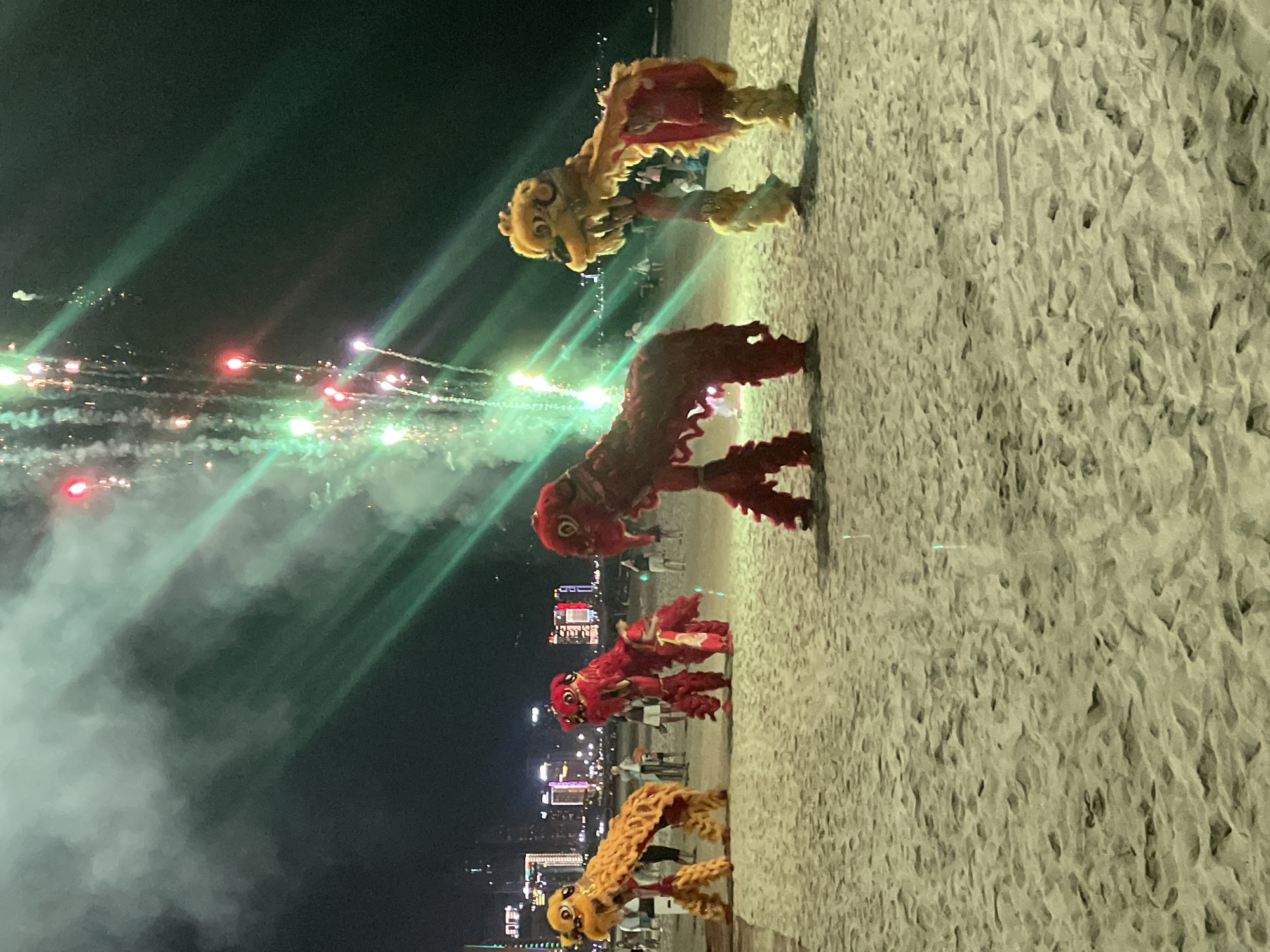 The image depicts a nighttime scene on a sandy beach in Da Nang, Vietnam. In the foreground, four performers are engaged in a lion dance, with two lions in red, one in yellow, and another in yellow and red. The lions are elaborate costumes with distinct heads and flowing bodies, supported by the performers underneath. Behind the lion dancers, a group of spectators is visible, casually dressed and observing the performance. The background features a cityscape with illuminated buildings and a dark sky, punctuated by a vibrant fireworks display. Green and red streaks of light from the fireworks cut across the frame, illuminating the smoke and the night sky. The sand on the beach is textured with footprints and small indentations.