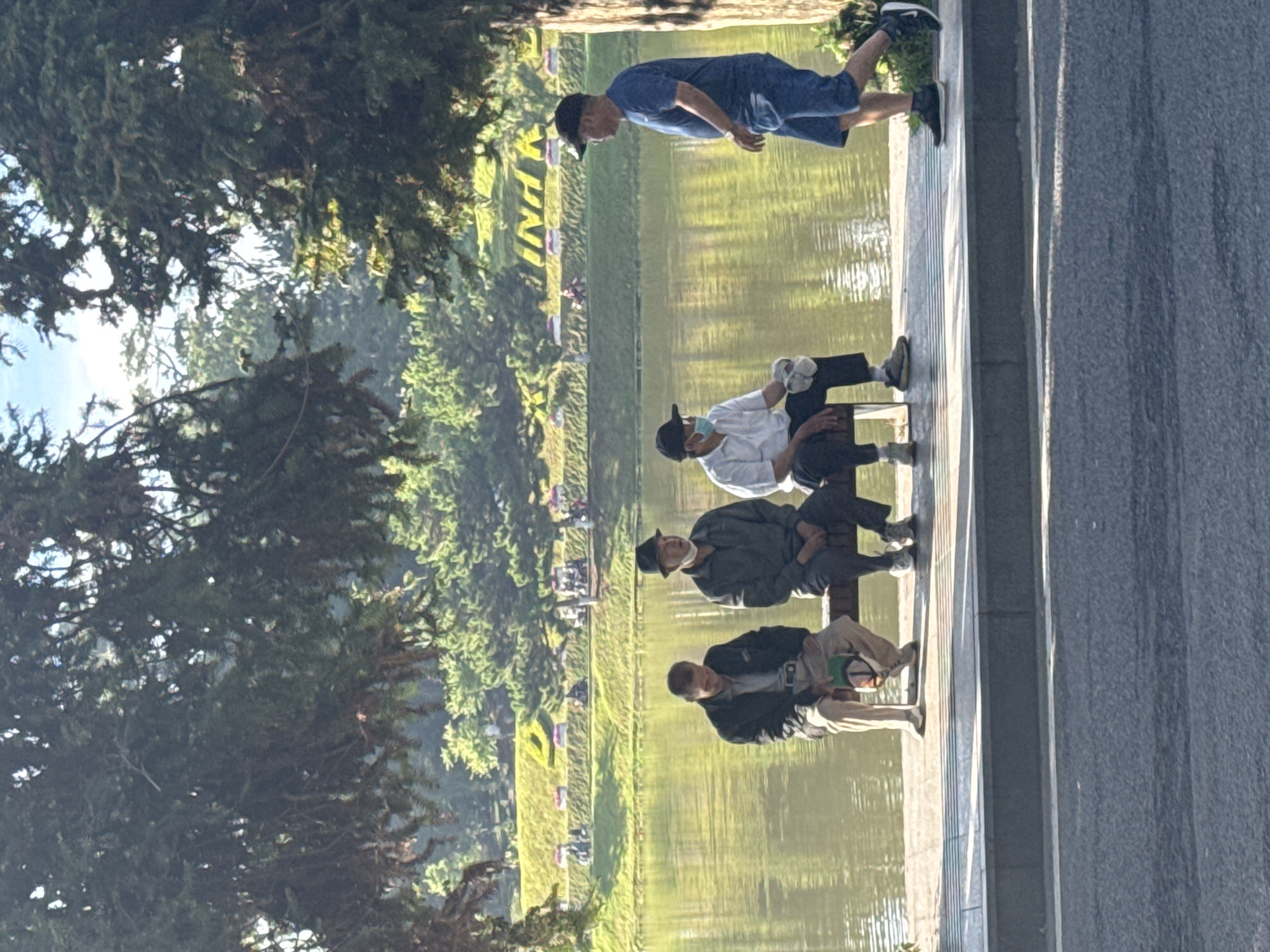 The image shows a group of four men sitting on a bench alongside a body of water in Da Lat, Vietnam. The men appear to be middle-aged, dressed in casual attire, and are oriented towards the water. One man is standing. The backdrop includes greenery, trees, and other vegetation, indicating an outdoor setting, likely a park or public space near a lake. The water is calm. There is a walkway next to the bench made of concrete. The overall scene suggests a tranquil and relaxed atmosphere.