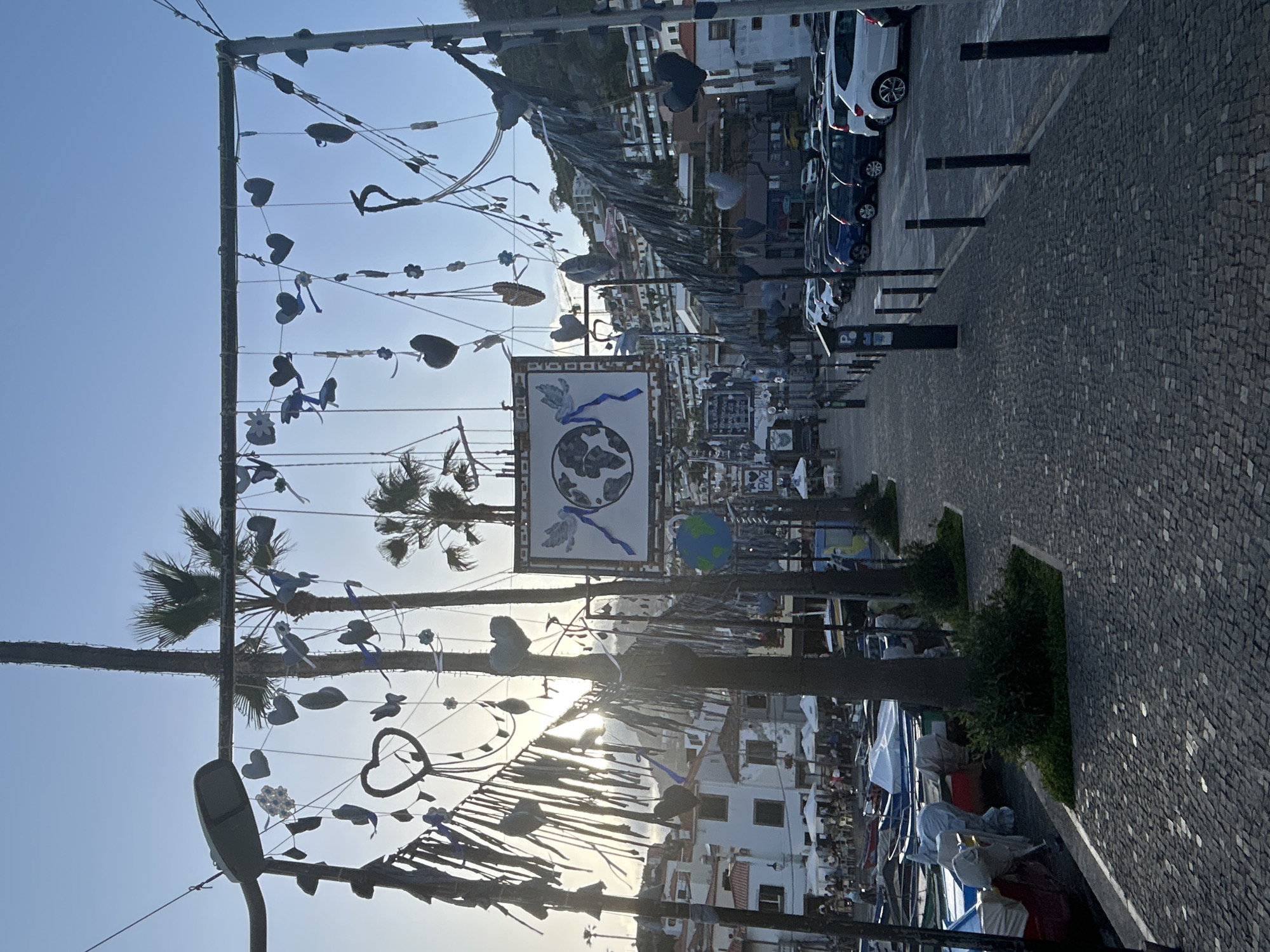 The image captures a street in Câmara de Lobos, Portugal, decorated with strung hearts, flowers, and birds suspended above a walkway. A sign depicting a globe and a dove is centered among the decorations. The walkway is paved with cobblestones, lined with plants, and features several modern bollards. Cars are parked parallel to the walkway. Buildings, some partially obscured by palm trees, stand in the background. People are visible in the distance on the harbor, near boats. The sky is a pale blue with the sun setting at the end of the street.
