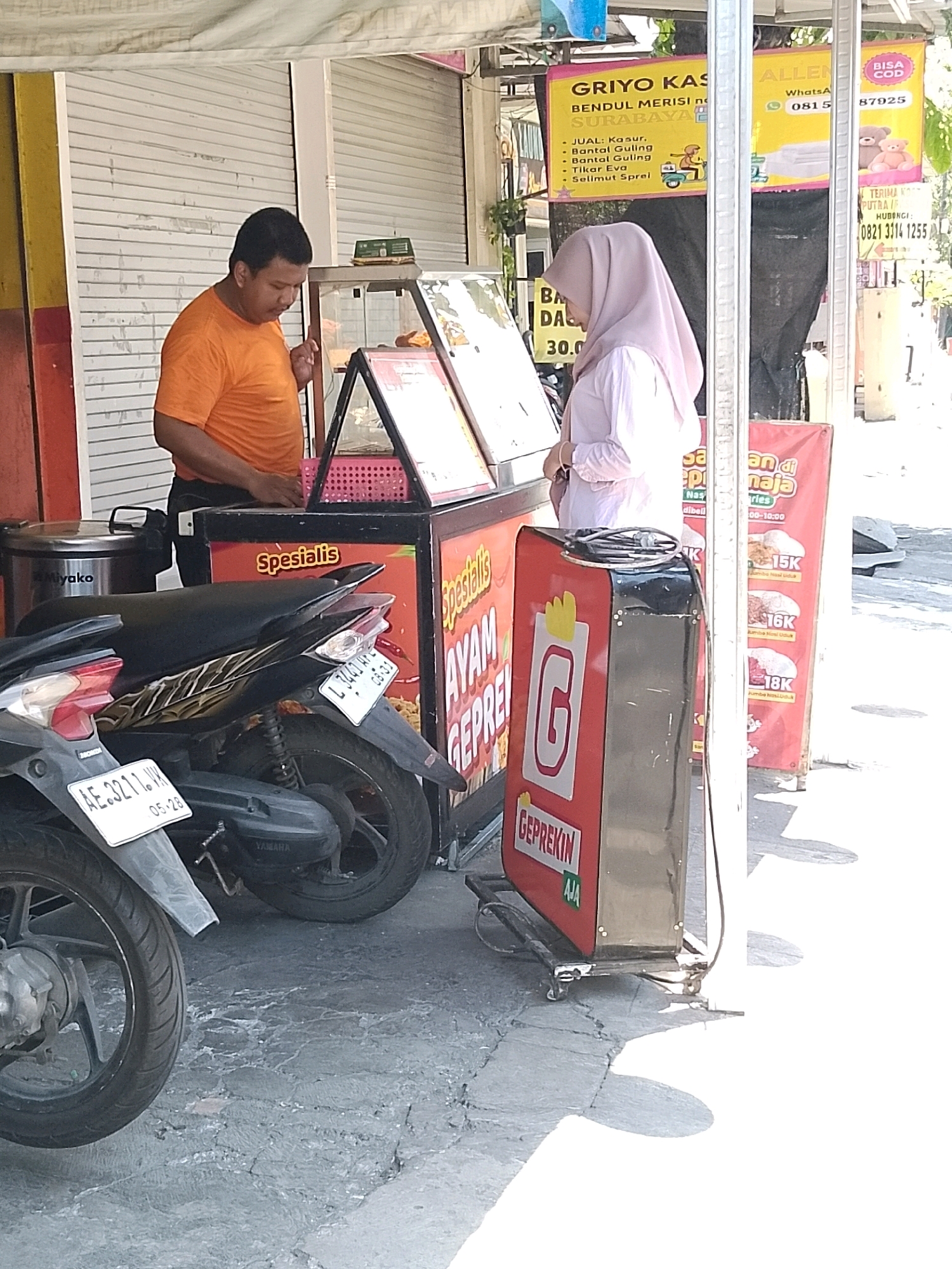 A medium shot captures an outdoor street food stall in Surabaya, Indonesia, on a sunny day. A male stall owner, wearing an orange t-shirt, stands behind a counter preparing food. To his left, a female customer in a light pink hijab is facing the counter, looking down. In front of the stall, two motorcycles are parked. The stall itself is adorned with various food advertisements and signage. One banner, in yellow, reads "ALLEN" and displays a WhatsApp number, "0815 87925". Another banner, predominantly pink, advertises "GRIYO KAS" and lists items such as "JUAL: Kasur, Bantal Guling, Tilam Eva, Selimut Sprei". A red sign on the stall reads "Spesialis AYAM GEPREK". The ground is paved with cracked concrete. The overall scene depicts a bustling commercial transaction in a typical Indonesian urban setting.