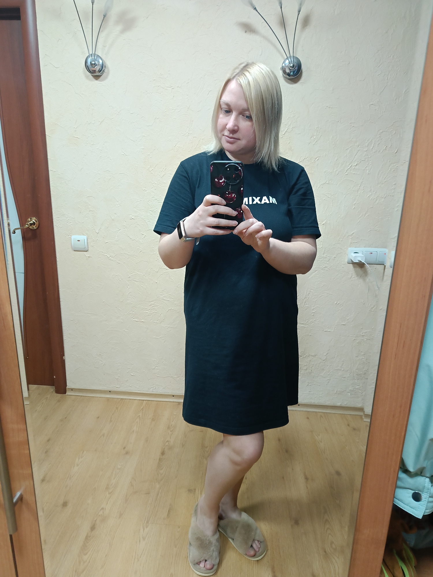 A woman with short blonde hair is standing in front of a mirror, taking a selfie. She is wearing a black t-shirt dress with white lettering and fuzzy slippers. She is holding a smartphone with a red cherry-themed case. The background includes a beige wall with light fixtures and electrical outlets. The floor is wooden. The scene appears to be inside a home. Given the prompt's mention of Chaikovsky, Russia, it is assumed that this is the location.
