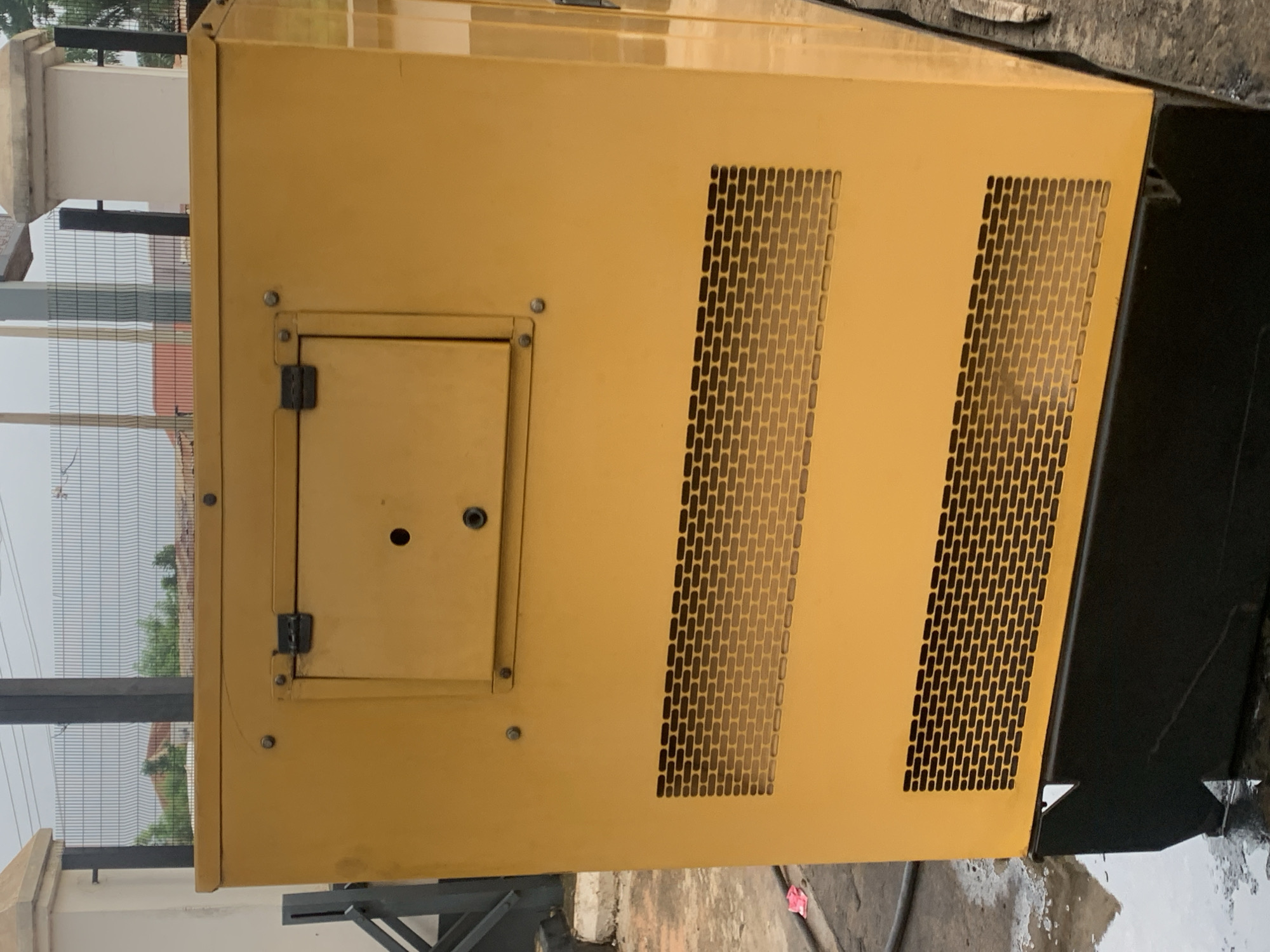 An exterior medium shot shows the side and back of a large, yellow generator. The generator is situated outdoors on a wet, paved surface. The generator is a rectangular prism with vents on its side and a smaller access panel on the back. The access panel has two black clasps and a circular hole in its center. The vents are rectangular and have a brick-like pattern. The generator rests on a black base. In the background, a metal fence with a diamond pattern encloses an area with trees and buildings. The sky is overcast and cloudy.