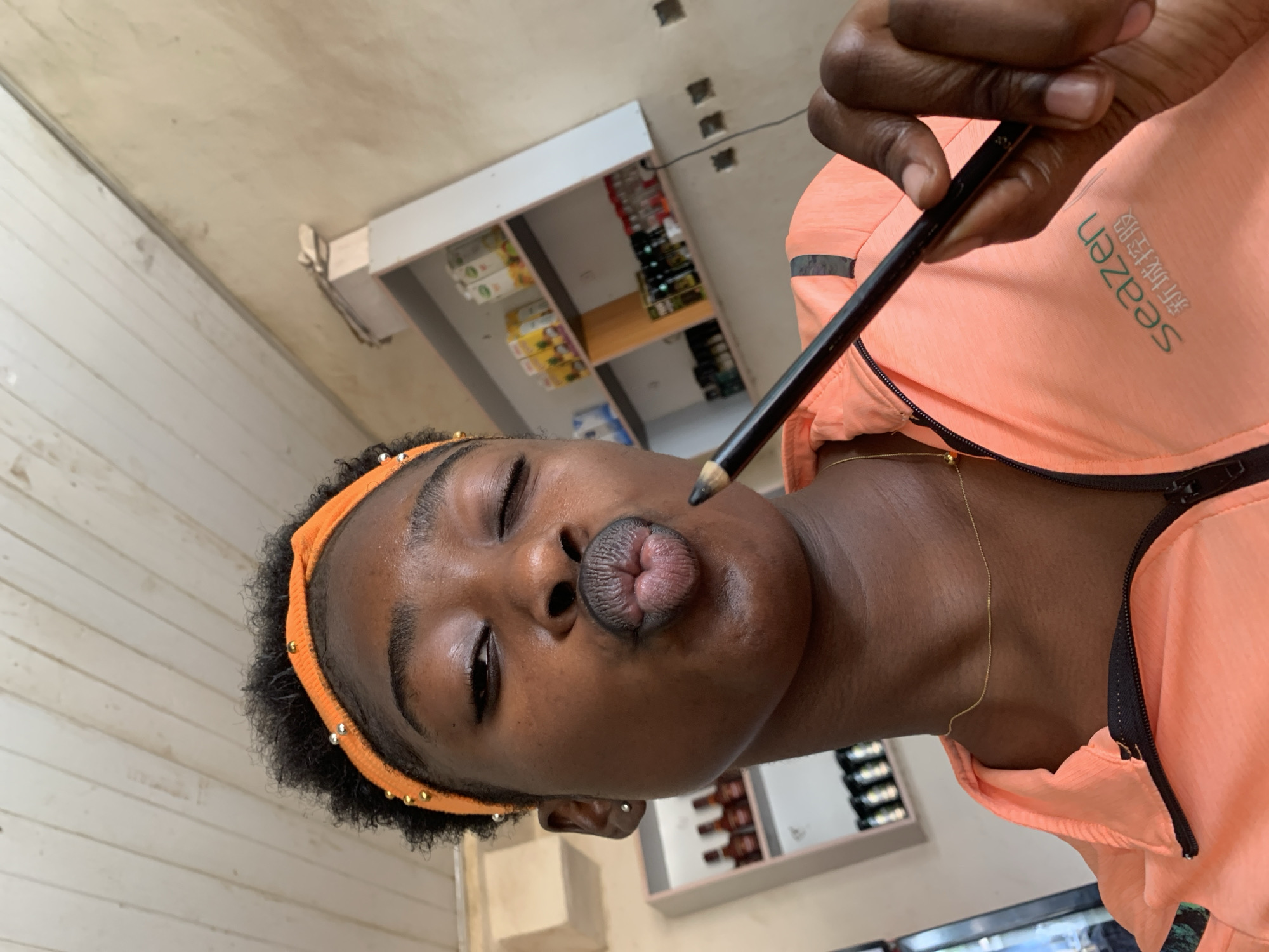 A person with dark skin and wearing an orange headband with gold accents is holding a black pencil near their face. They are wearing an orange athletic jacket with white lettering and a black zipper. A thin gold chain necklace is visible on their neck. They are pursing their lips. The background includes a wall with horizontal wooden planks and shelves stocked with various bottled products. The location is New Stadium, Nigeria.
