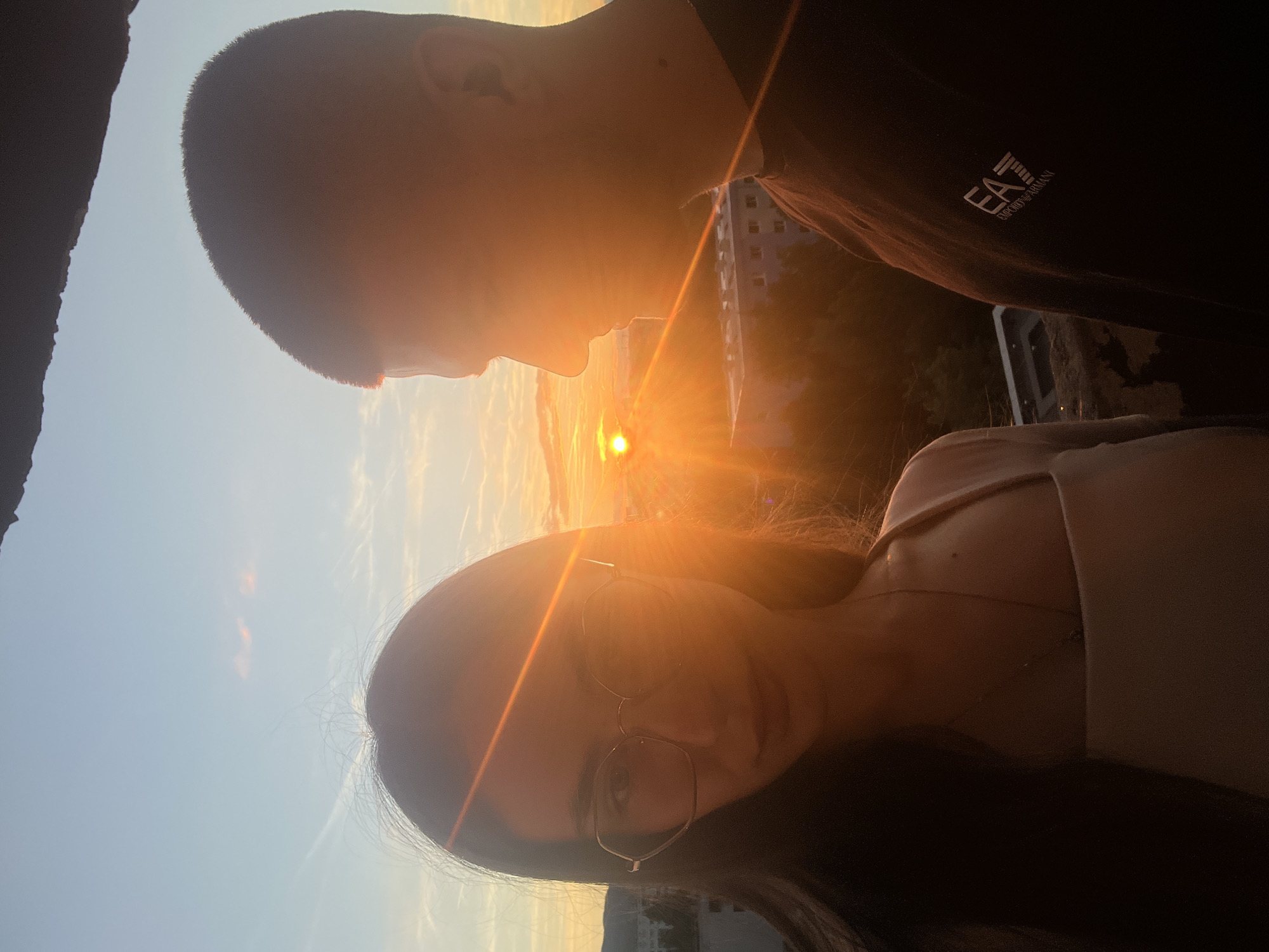 A medium shot captures two individuals, a woman and a man, silhouetted against a vibrant sunset over a city. The woman is positioned on the left, her face turned towards the viewer, wearing glasses and a light-colored top. Her dark hair is visible on the left side of the frame, and she is looking towards the camera. To her right, the man is shown in profile, facing away from the camera and towards the sunset. He is wearing a dark t-shirt with a visible logo on the right side of his chest. The sunset is the focal point, with the sun low on the horizon, casting a warm, orange glow across the sky and illuminating the scene. In the background, city buildings and trees are visible, appearing muted by the light of the setting sun. The overall impression is one of a serene and romantic moment shared by the couple.