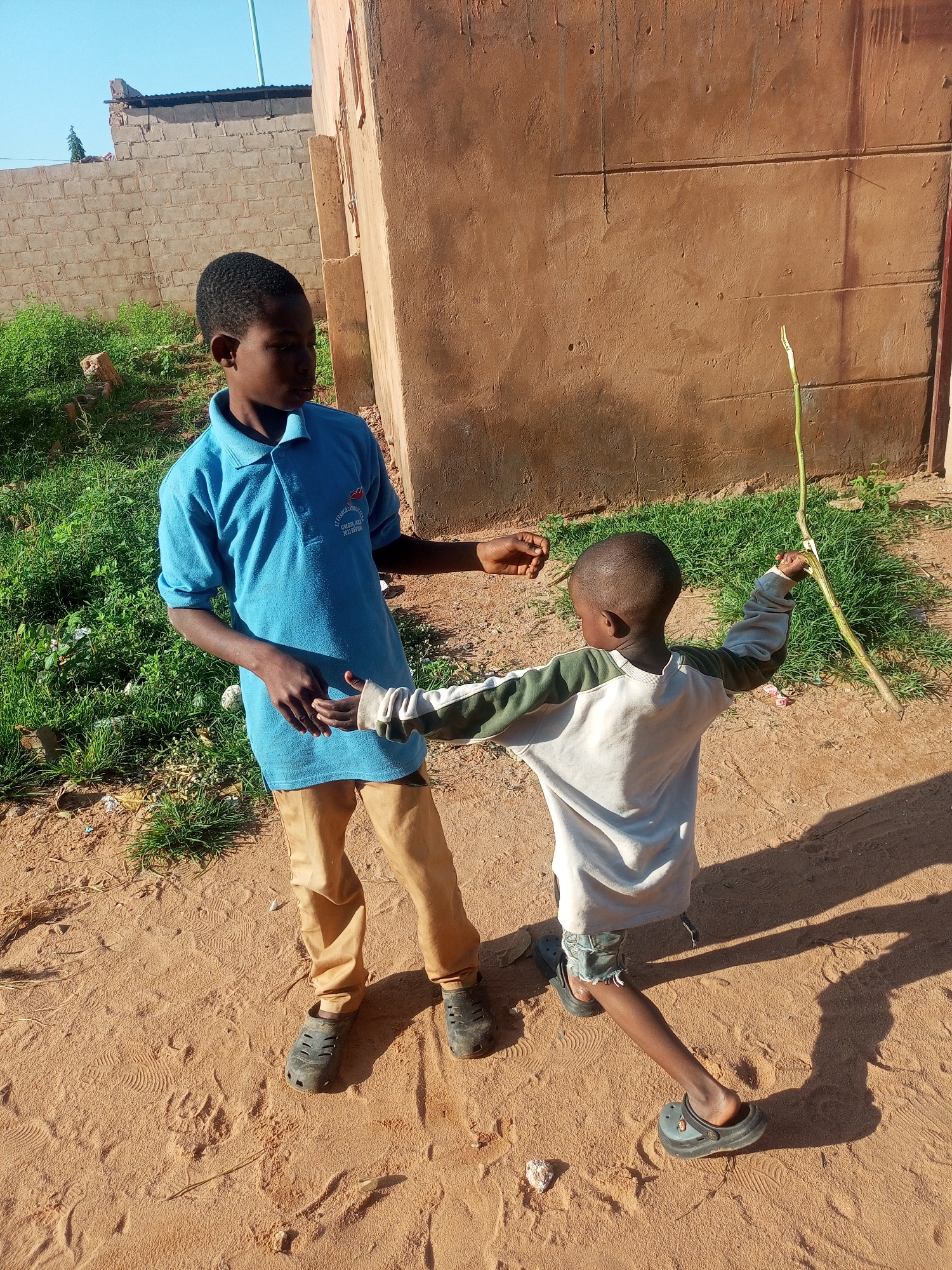 In Tempure, Nigeria, two young boys are captured in a bright, sunlit outdoor scene. The elder boy, wearing a blue polo shirt and tan pants, stands with his left arm extended towards the other child. The younger boy, dressed in a white and green long-sleeved shirt and shorts, is facing away from the camera, his arms outstretched as if interacting with the elder. He holds a long, thin green plant stalk in his right hand. They are standing on a sandy ground with some sparse green vegetation and debris scattered around. Behind them, a rustic, ochre-colored building with a corrugated metal roof is visible, and to the left, a textured brick wall extends into the background. The lighting suggests it is daytime, with clear blue skies overhead.