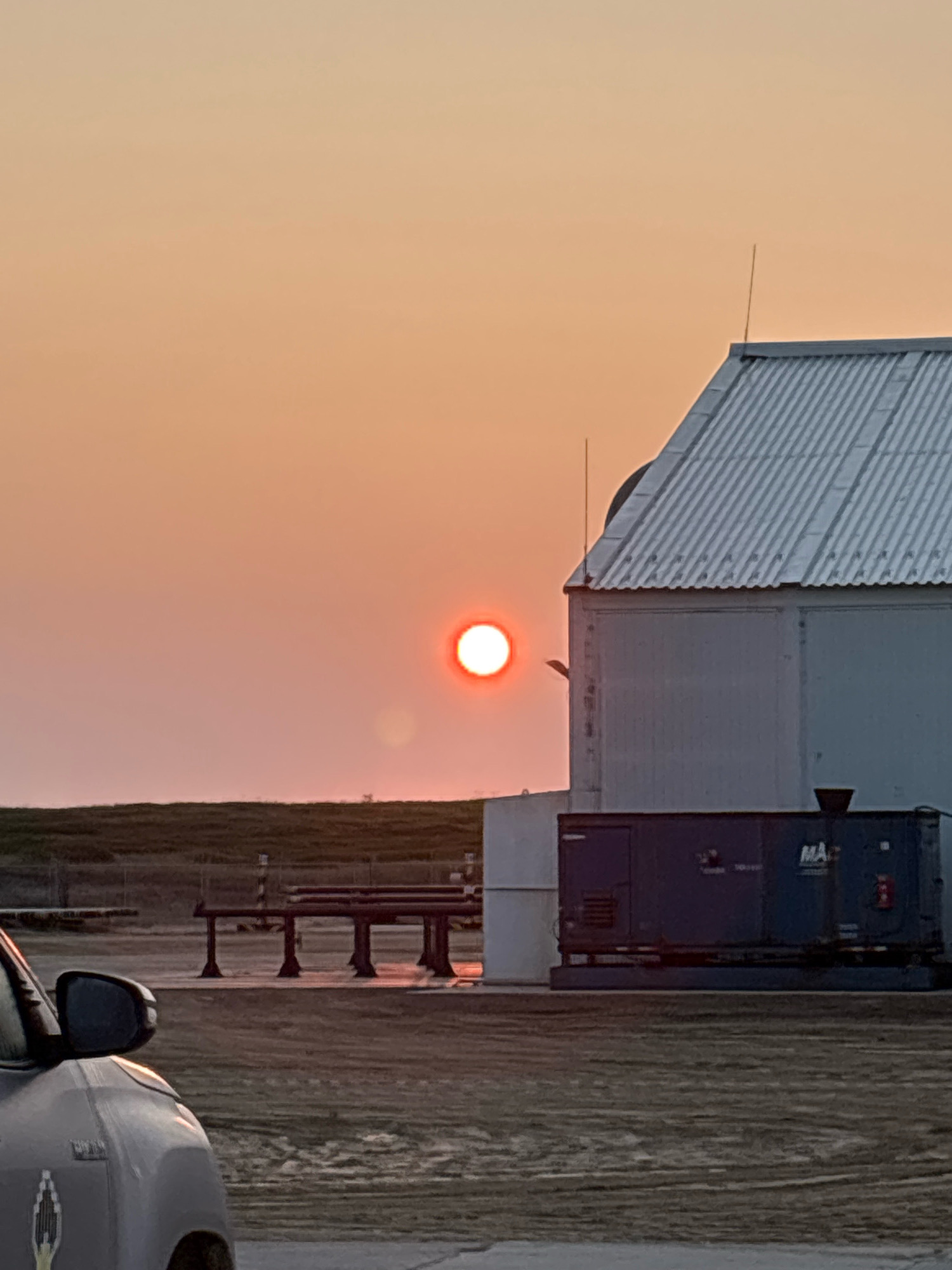 The image shows a sunset scene in a possibly industrial or rural location. The sun, a bright orb with a reddish glow, is visible in the sky, casting a warm light. A portion of a white building with a corrugated metal roof is on the right side of the frame. In front of this building, there is a dark blue industrial generator with "MA" and other markings visible. Further back, a dark-colored car is partially visible in the lower left corner. The ground appears to be dirt or gravel, with some sparse vegetation in the background. There are also some metal structures and poles in the mid-ground. The sky is a gradient of orange and yellow, typical of a sunset. There are no people visible in the image. The location is described as Unknown, Russia, but there are no specific landmarks or identifying features to confirm this.
