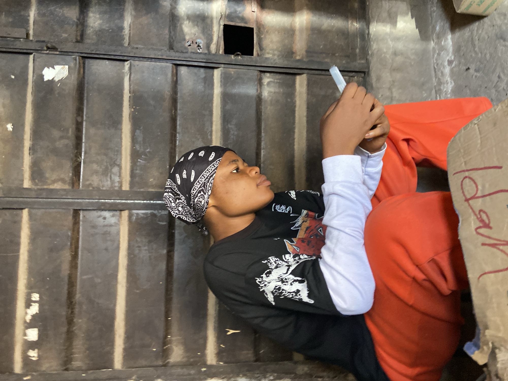 A young person, wearing a black bandana with a white paisley print, a black long-sleeved t-shirt with white graphics, and orange sweatpants, is seated and looking down at a mobile phone held in their hands. The person is positioned in front of a dark metal, corrugated structure, possibly a door or wall, that appears weathered and somewhat worn. The ground in front of the person is concrete and shows some debris. The scene is captured outdoors in the city of Ogidi, Nigeria, likely in a public or semi-public space, suggested by the urban setting and the presence of a cardboard box with writing on it in the foreground. The lighting suggests it is daytime.