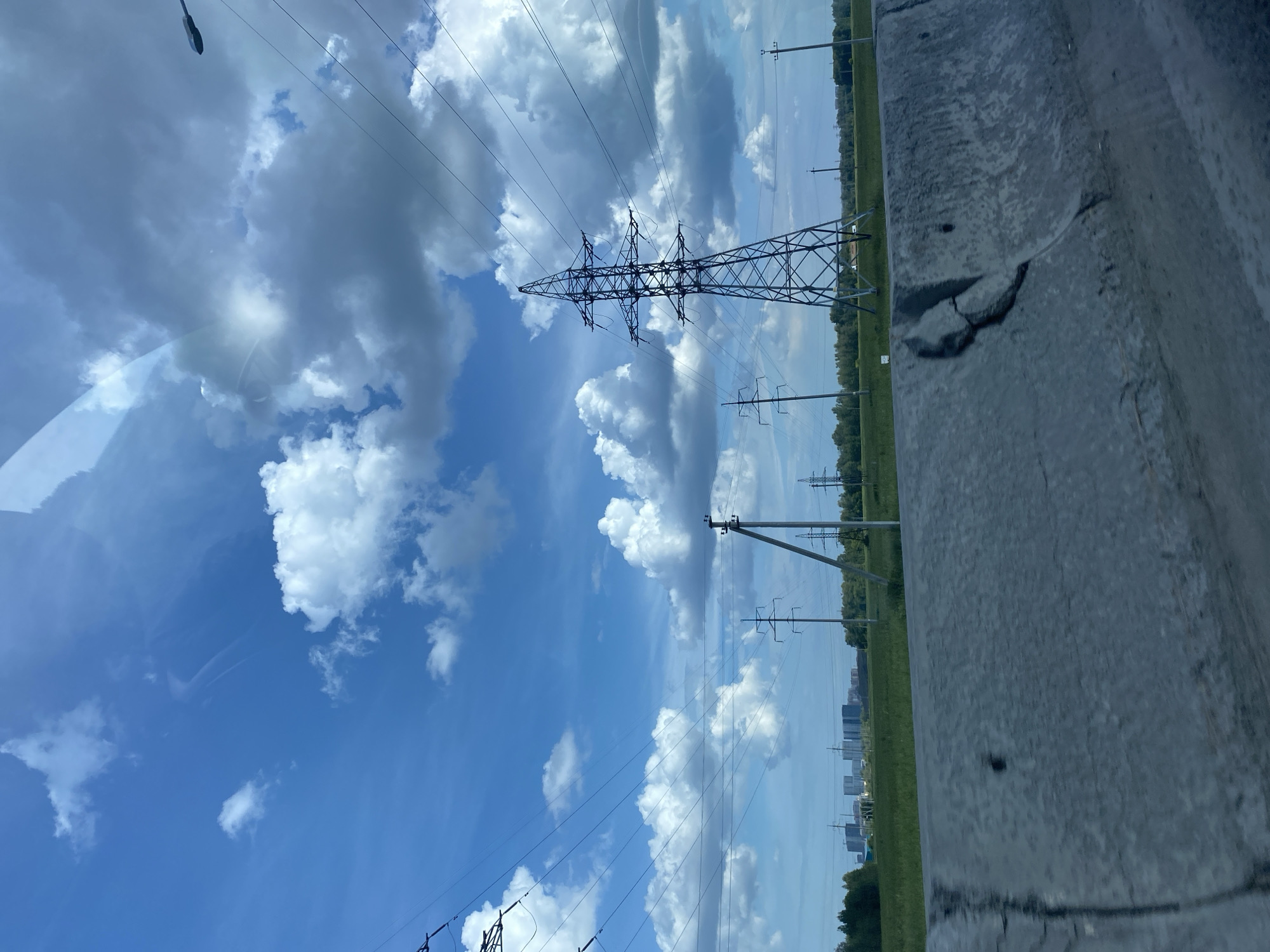 The image depicts a wide, slightly hazy daytime view of a landscape in Tyumen, Russia, dominated by a bright blue sky filled with fluffy white and grey clouds. In the foreground, a rough, weathered concrete barrier with visible cracks runs across the lower portion of the frame. Beyond the barrier, a green grassy field stretches to the horizon. Several power lines crisscross the sky, supported by tall, metal lattice towers and shorter concrete poles. One prominent, large metal tower stands to the right of the frame. In the far distance, a cluster of modern, light-colored buildings forms a city skyline. The overall impression is that of a semi-urban or suburban environment with industrial infrastructure set against a natural backdrop.