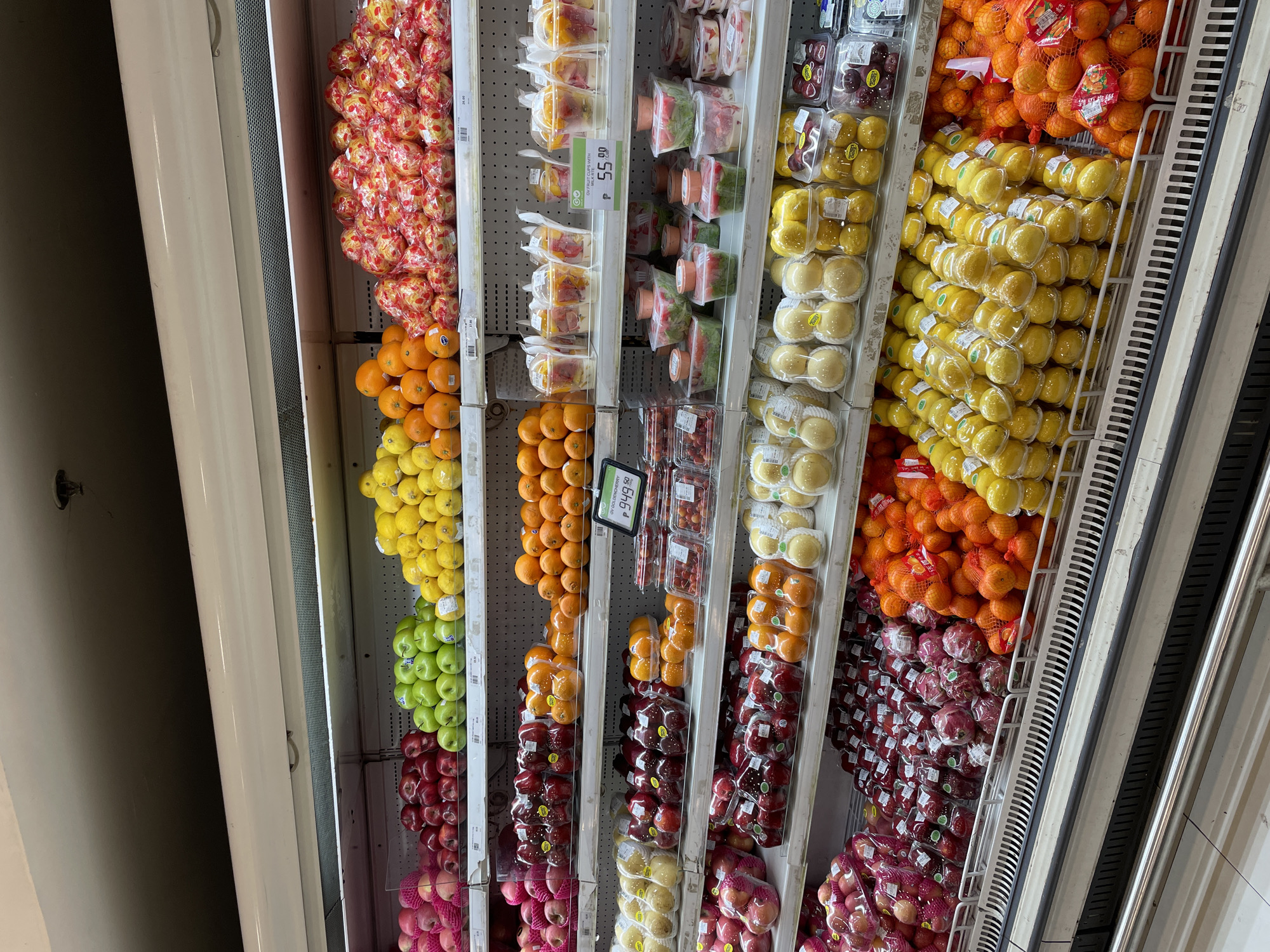 This image depicts a well-stocked produce section of a supermarket, likely located in Davao City, Philippines, given the presence of a price tag in Philippine Pesos (₱949.50) and a price of ₱55.00 with the currency symbol displayed. The shelves are organized with various fruits, including apples, oranges, lemons, and what appear to be pears or similar fruits, as well as pre-packaged fruit cups and possibly salads or other prepared food items. The lighting is bright and fluorescent, typical of a retail environment. No people are visible in the image. The overall scene suggests a typical day in a grocery store with fresh produce for sale.