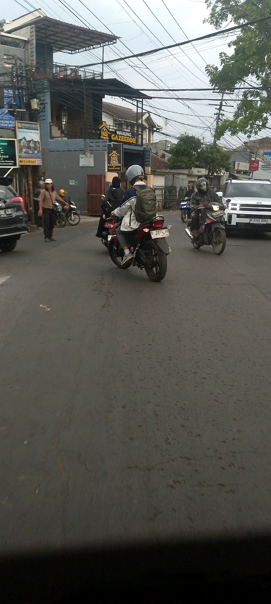 The image captures a street scene in Bandung City, Indonesia, likely during the daytime due to the ambient light. The focus is on a road with several vehicles and pedestrians. On the left, a dark car is parked, and further back, a man in a light brown shirt and dark pants is walking. Several motorcycles are also present: one is red with two riders, one is blue with one rider, and another is a darker color with a single rider, all navigating the asphalt road. In the background, buildings are visible, including one with a prominent sign that reads "WARUNG GAZEEBOO," suggesting it is a food stall or small restaurant. Overhead, a complex network of electrical wires crisscrosses the sky, indicating an urban environment. The road surface shows signs of wear and tear, with cracks and patches visible. The overall impression is a snapshot of everyday urban life in Indonesia.