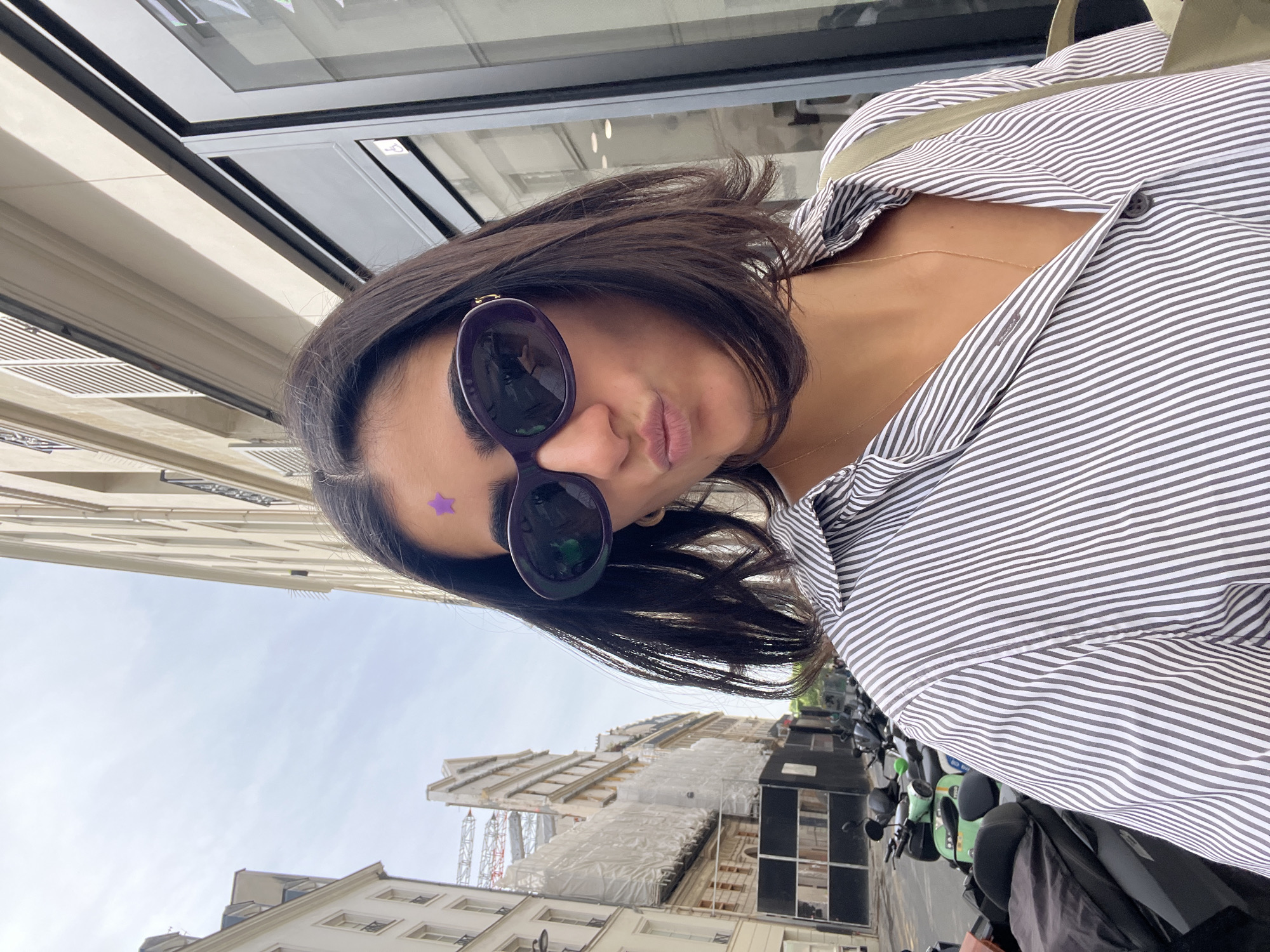 A woman with dark hair and sunglasses is posing in an urban environment. She is wearing a striped shirt and has a purple star on her forehead. The background shows buildings and scooters parked on the street. The location appears to be Paris, France, due to the architectural style of the buildings and street layout.