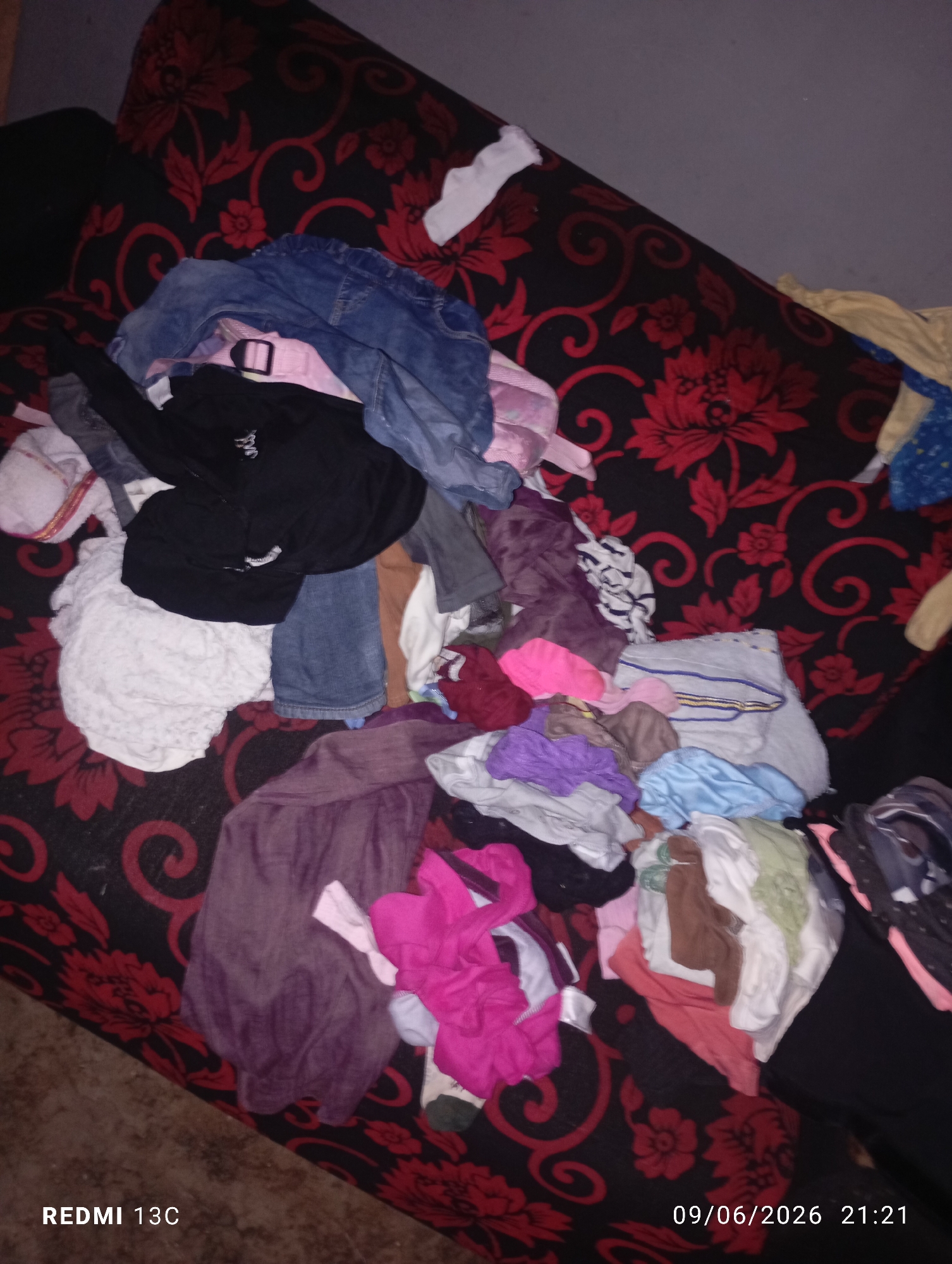 A pile of assorted clothing is scattered across a patterned sofa. The clothing includes a pair of blue jeans, a black jacket, a white towel, and various colorful garments like pink, purple, and blue items. The sofa has a dark background with a red floral pattern. The scene appears to be indoors, likely a domestic setting.