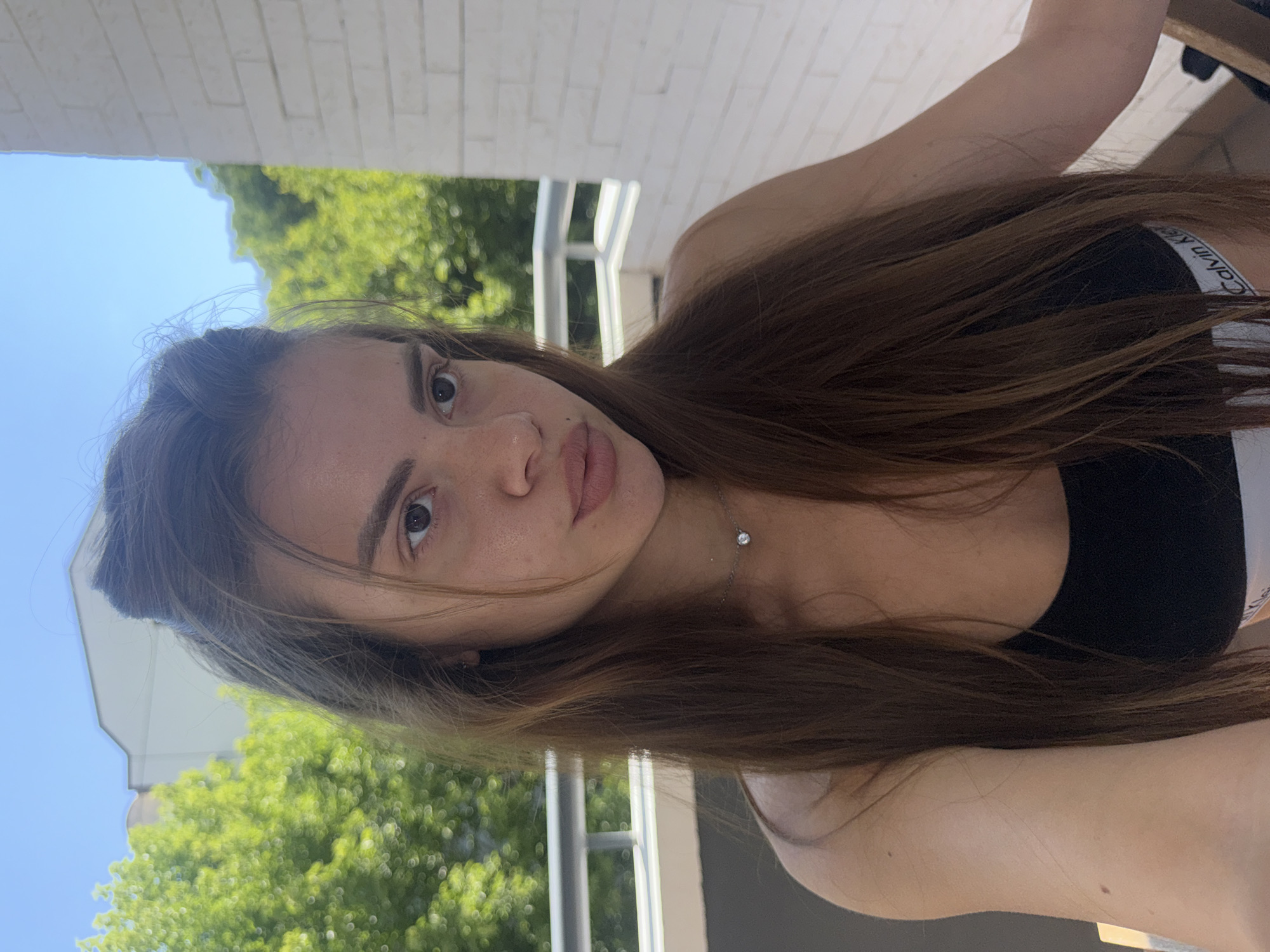 A young woman with long, brown hair is pictured from the chest up, looking towards the camera. She is wearing a black sports bra with a white band that has "Calvin Klein" printed on it. A small, silver pendant with a clear stone hangs around her neck. Her hair is partially pulled up, with some strands falling around her face. Her skin appears to have a light tan, and she has a small mole on her cheekbone.
The background suggests an outdoor setting, possibly a balcony or terrace. Green foliage from trees is visible behind her, along with a bright blue sky. To the right, a white brick wall and a white railing are present. The overall lighting indicates it is daytime. The scene does not provide any specific visual cues to pinpoint its location within the city of Plovdiv, Bulgaria.
