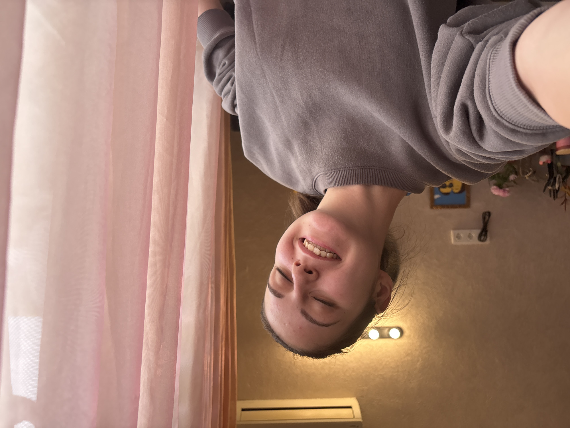 A young woman with light skin, wearing a gray sweatshirt, is smiling with her eyes closed. The image is rotated, placing the woman upside down. Behind her is a light brown wall, a set of vanity lights, and an air conditioning unit. A sheer pink curtain is to her left. A small, framed picture, a pink flower arrangement, and other decorative items are partially visible on a shelf behind her. The location of the image is Bulgaria, Bulgaria, but there is no way to verify the location.