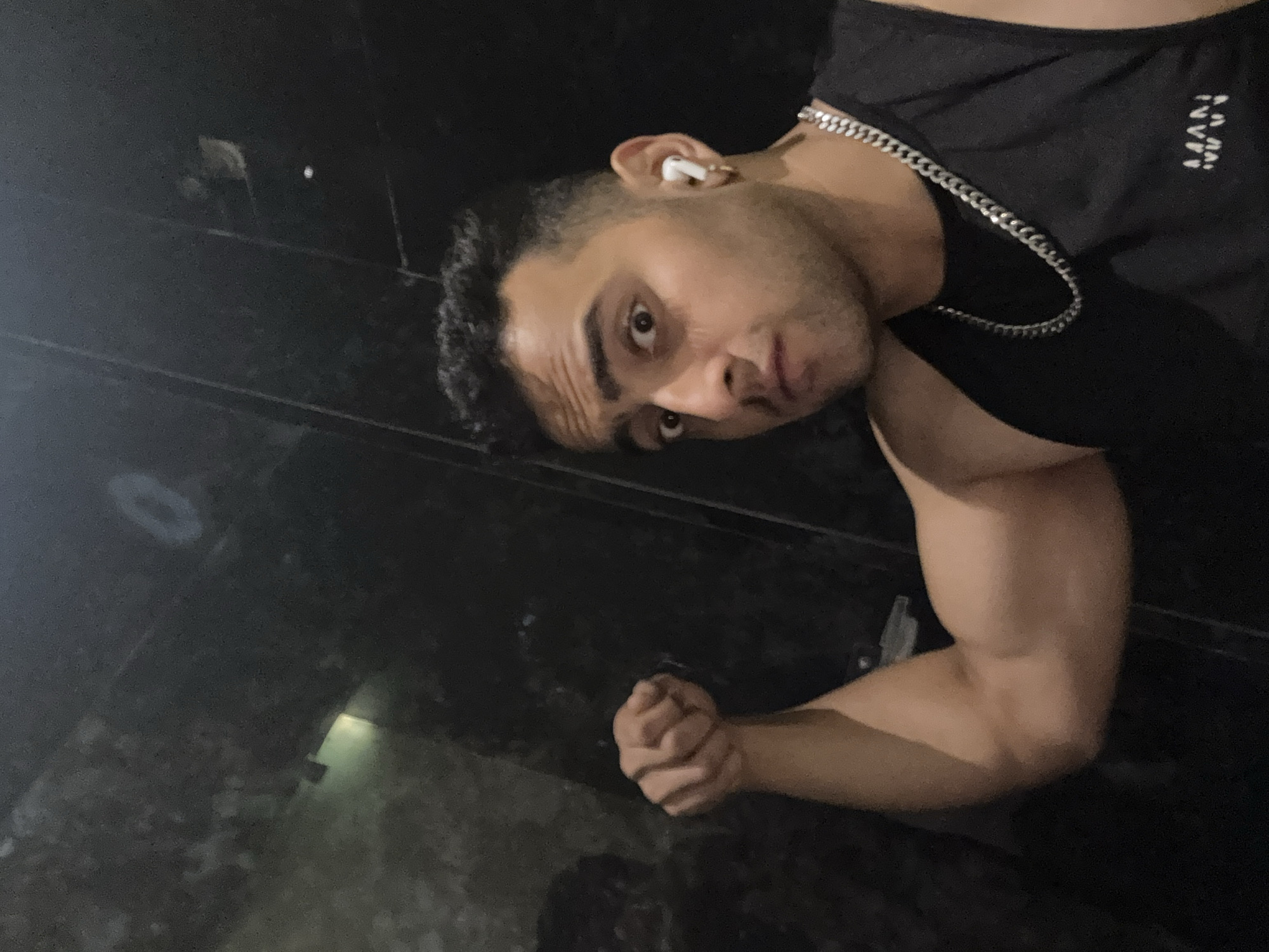 A man wearing a black tank top with the word "MAN" printed on it and a silver chain around his neck is posing for the camera. He is indoors and appears to be in a dimly lit environment, potentially a hallway or a garage, given the visible wall and floor textures. His right arm is flexed to showcase his bicep, and his left hand is clenched into a fist. He is wearing a white wireless earbud in his left ear and looking directly at the camera with a slightly intense expression. It is not possible to pinpoint the exact location in Kolkata, India, based solely on the visual information provided.