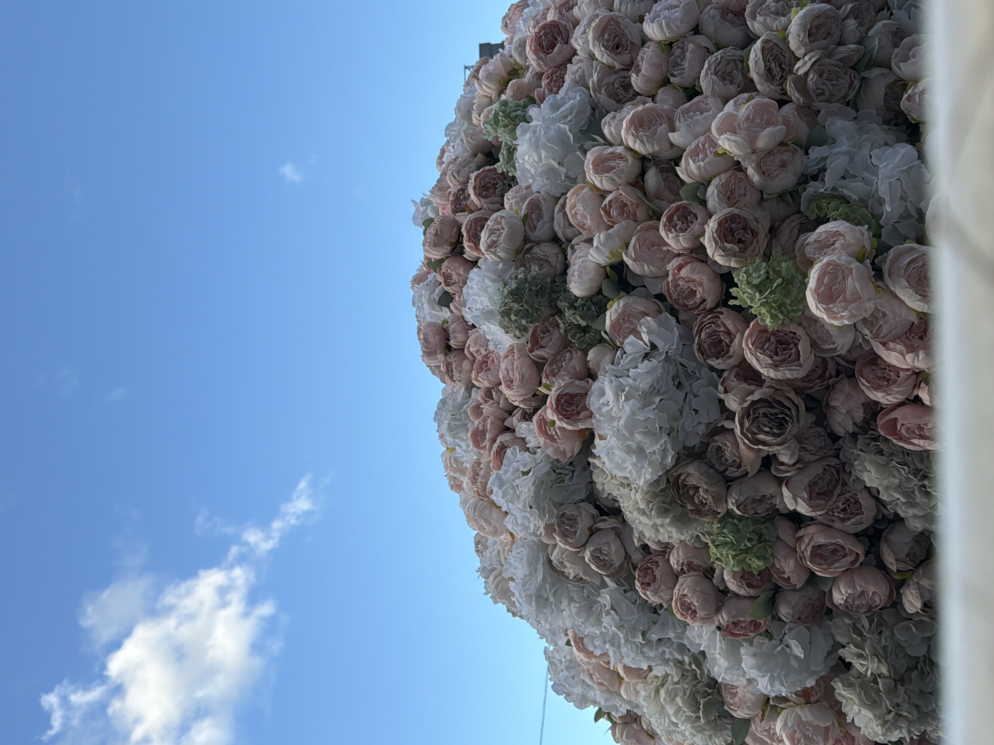 A large, spherical mound of artificial flowers, composed primarily of pink, white, and muted green peonies, is displayed outdoors under a blue sky with scattered white clouds. The flowers are densely packed, creating a textured and voluminous surface that obscures any underlying structure. The mound appears to be positioned on a raised platform, indicated by a white, blurred surface in the foreground. In the upper right corner of the image, a dark, rectangular object, possibly a building or a portion of a structure, is partially visible against the sky. The scene is devoid of any people or discernible activities.
