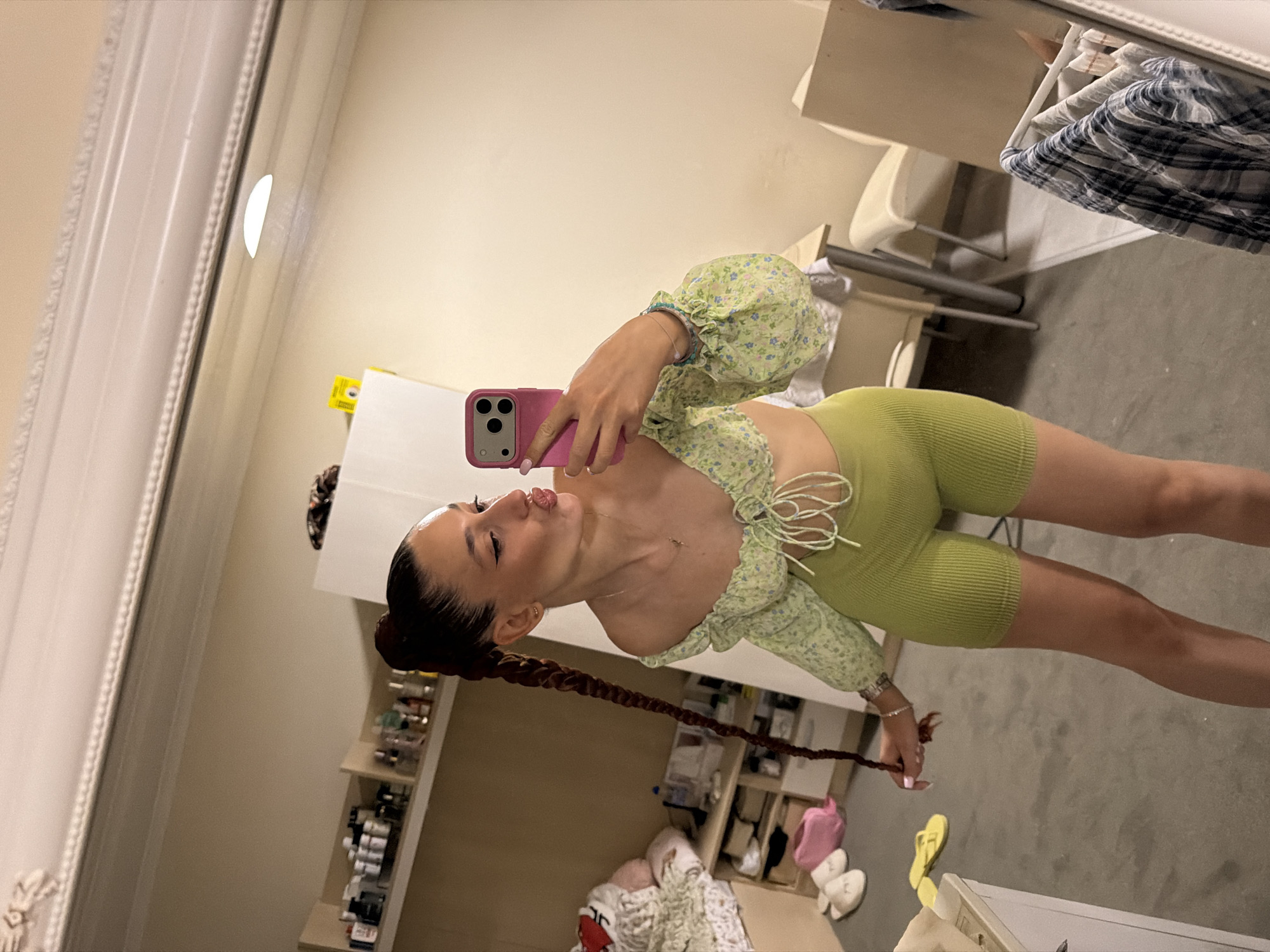 A young woman with dark hair in a long braid is taking a mirror selfie in what appears to be a bedroom. She is wearing a green floral off-the-shoulder crop top and lime green high-waisted biker shorts. She is holding a pink smartphone in her right hand and puckering her lips towards the mirror. Her left hand is holding her braid. In the background, there are shelves filled with various items, a bed with blankets and pillows, and a table with chairs. The scene is dimly lit, and there is a large mirror reflecting the room. The image does not provide any specific visual cues to indicate the location in Sofia, Bulgaria.