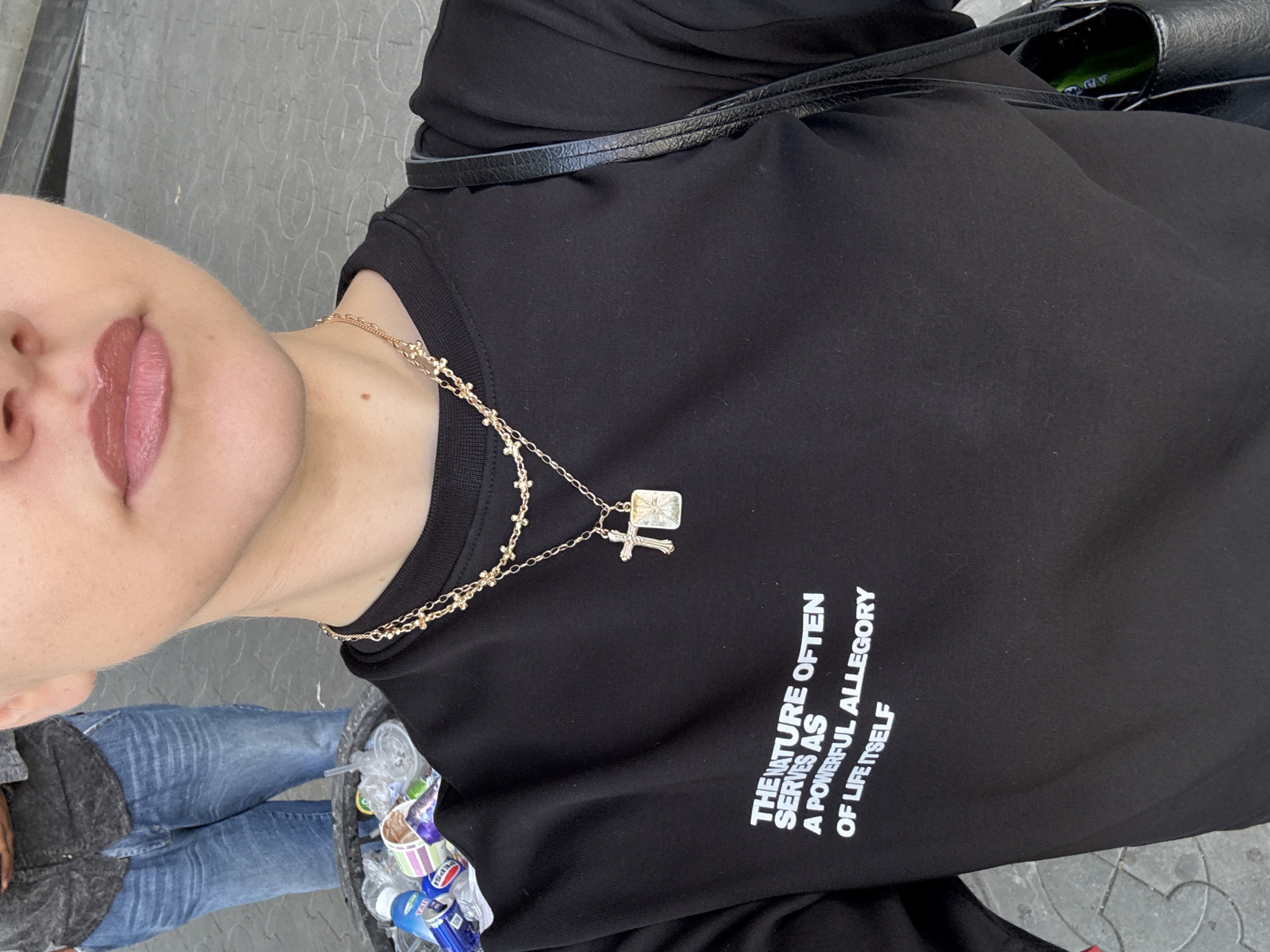 The image shows a close-up of a person wearing a black t-shirt with white text. The text on the t-shirt reads: "THE NATURE OFTEN SERVES AS A POWERFUL ALLEGORY OF LIFE ITSELF". The person is also wearing a gold necklace with a cross pendant. In the background, there is a trash can filled with plastic bottles and other items, and another person wearing jeans and a grey shirt. The ground appears to be paved with concrete or stone. The scene suggests an outdoor urban environment, possibly in Yerevan, Armenia, given the provided context.